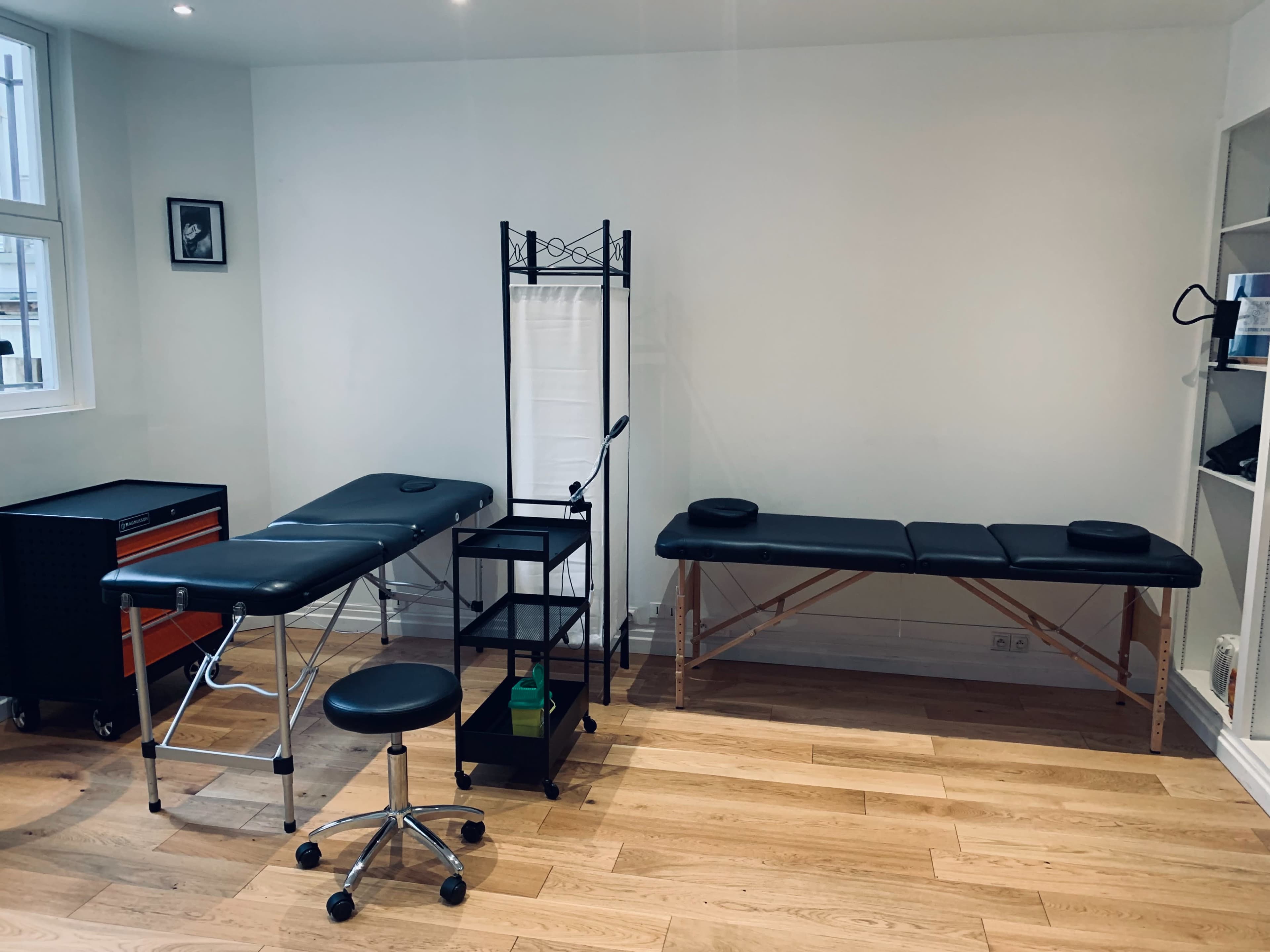 A treatment room with two therapy tables, a black rolling stool, and a storage unit against a light-colored wall.