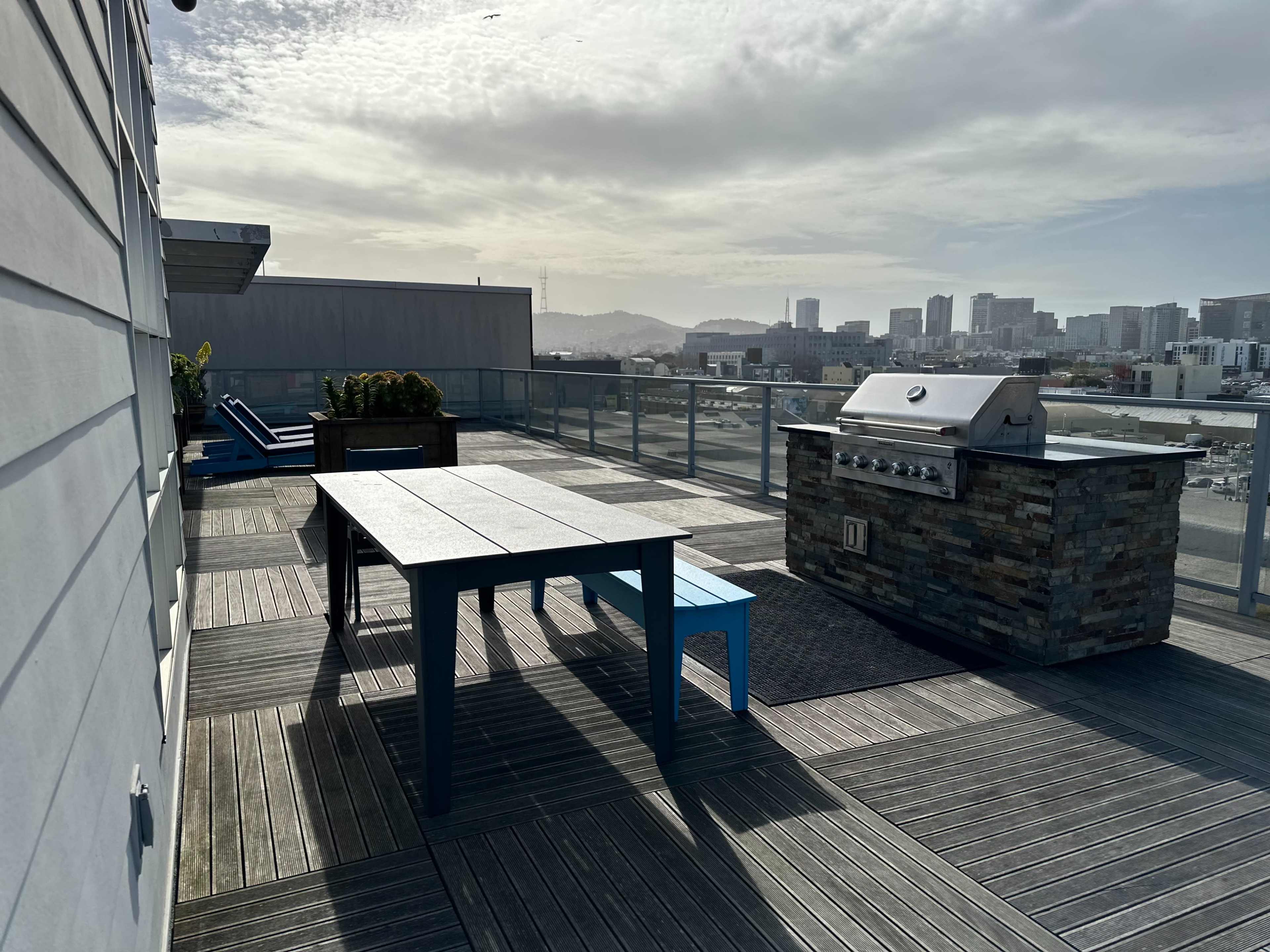 A rooftop patio with a large table, a blue chair, a stone barbecue grill, and a view of city buildings in the background.