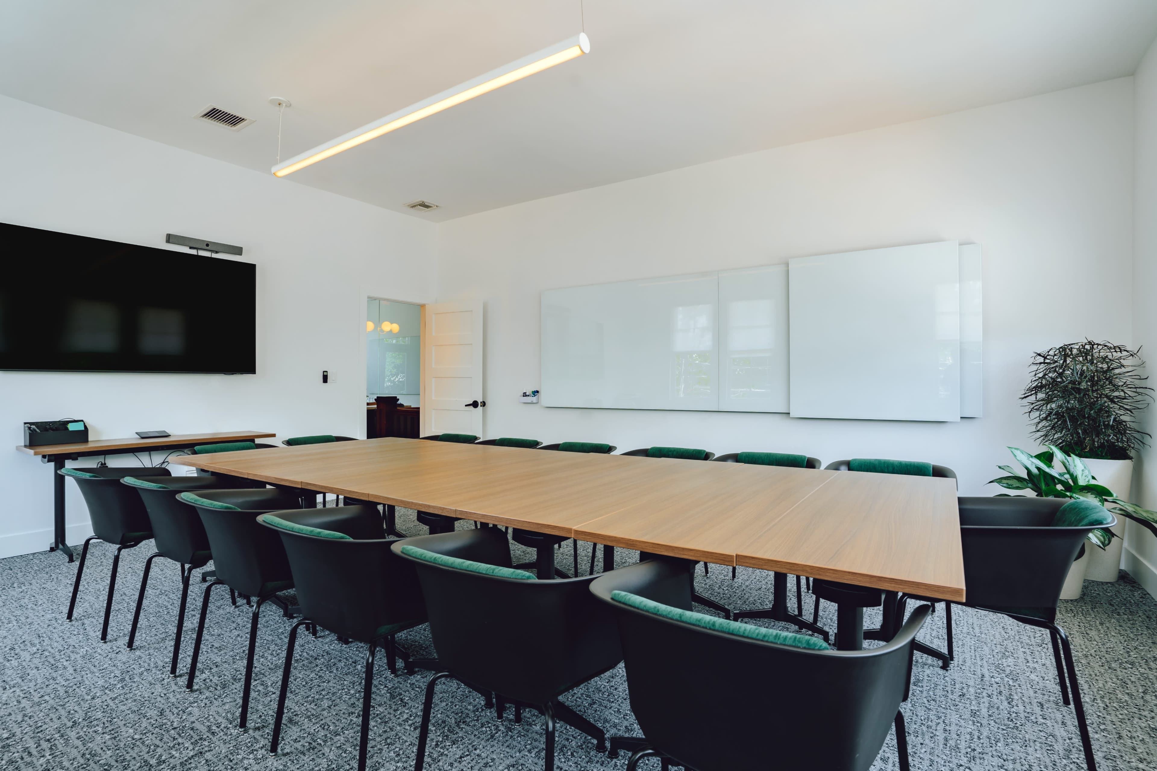 The image shows a modern conference room with a long wooden table surrounded by black chairs, a large screen on the wall, and whiteboards on the adjacent walls.