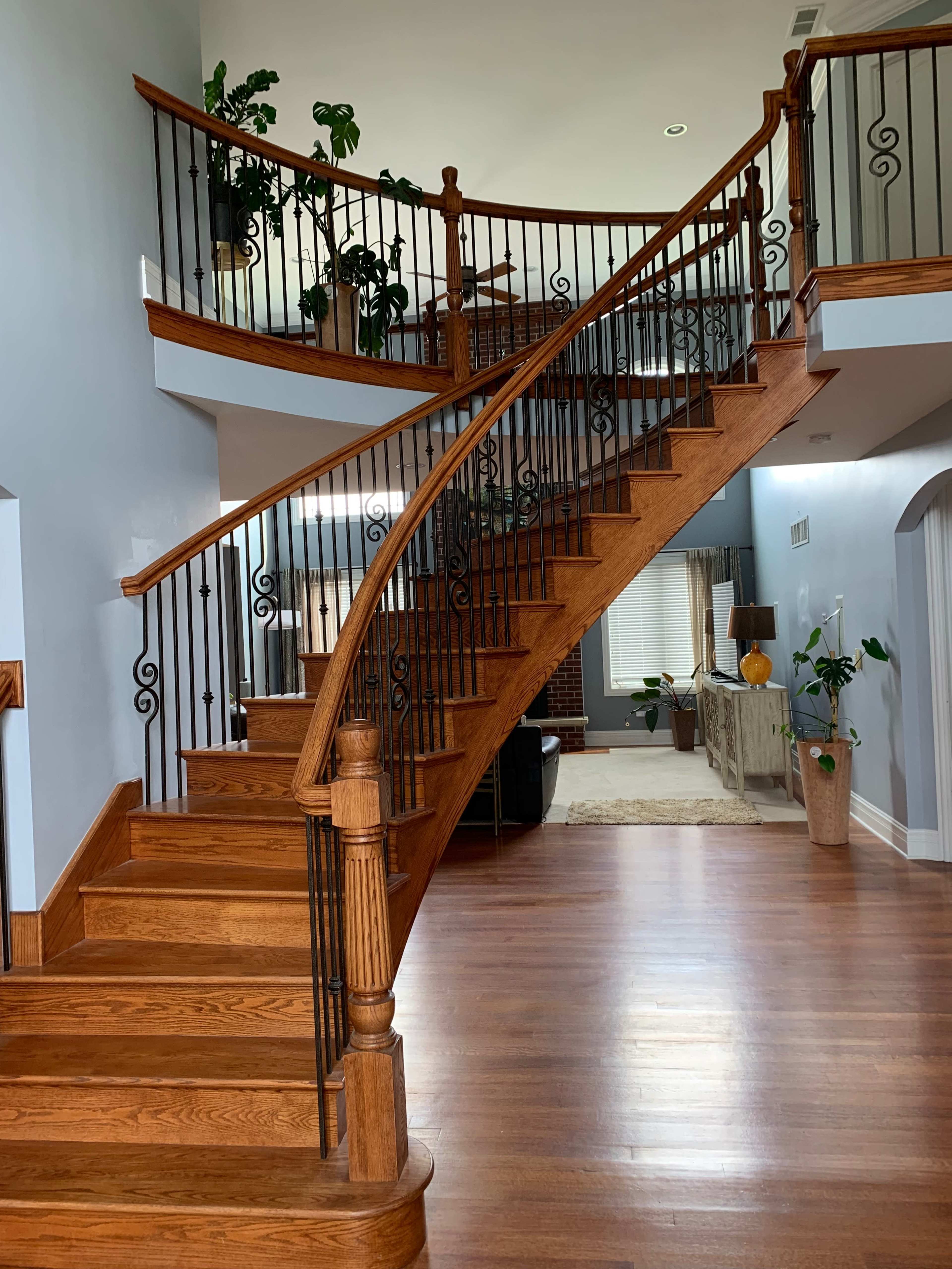 The image shows a wooden spiral staircase with wrought iron railing leading to an upper level in a well-lit foyer.
