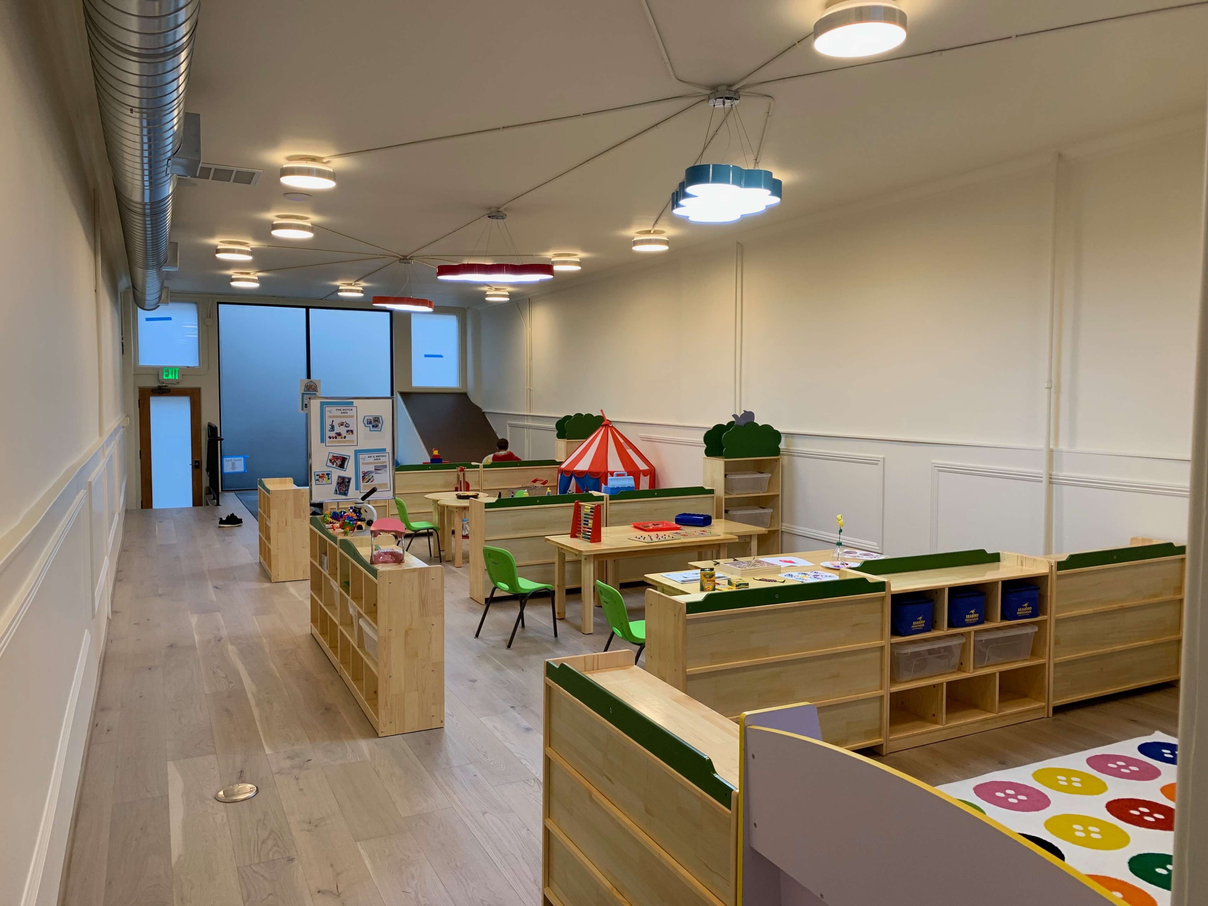 A brightly lit classroom with wooden play furniture, activity stations, and educational posters arranged throughout the space.