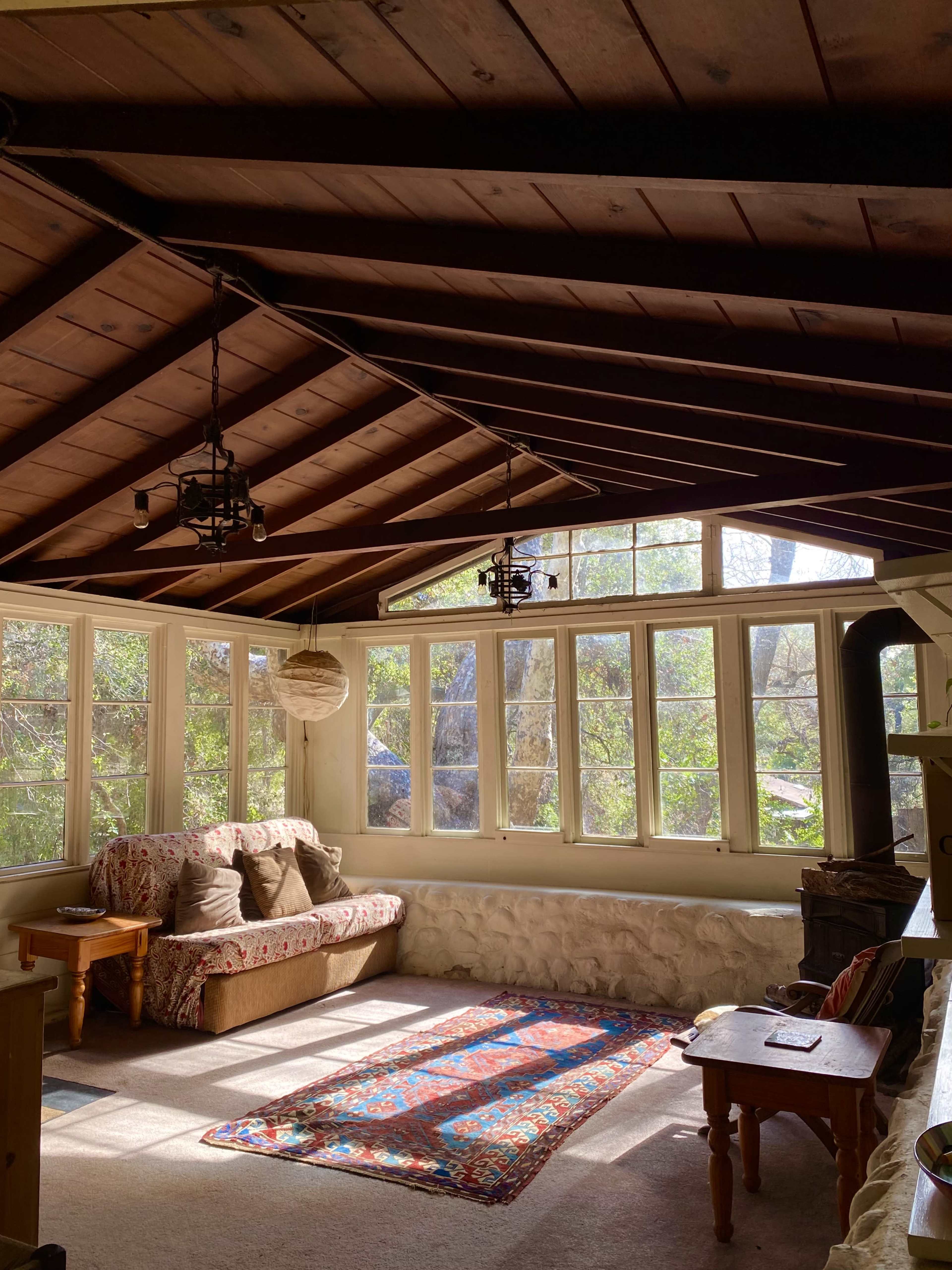 A sunlit interior features a cozy seating area with a patterned rug and large windows that overlook a natural landscape.