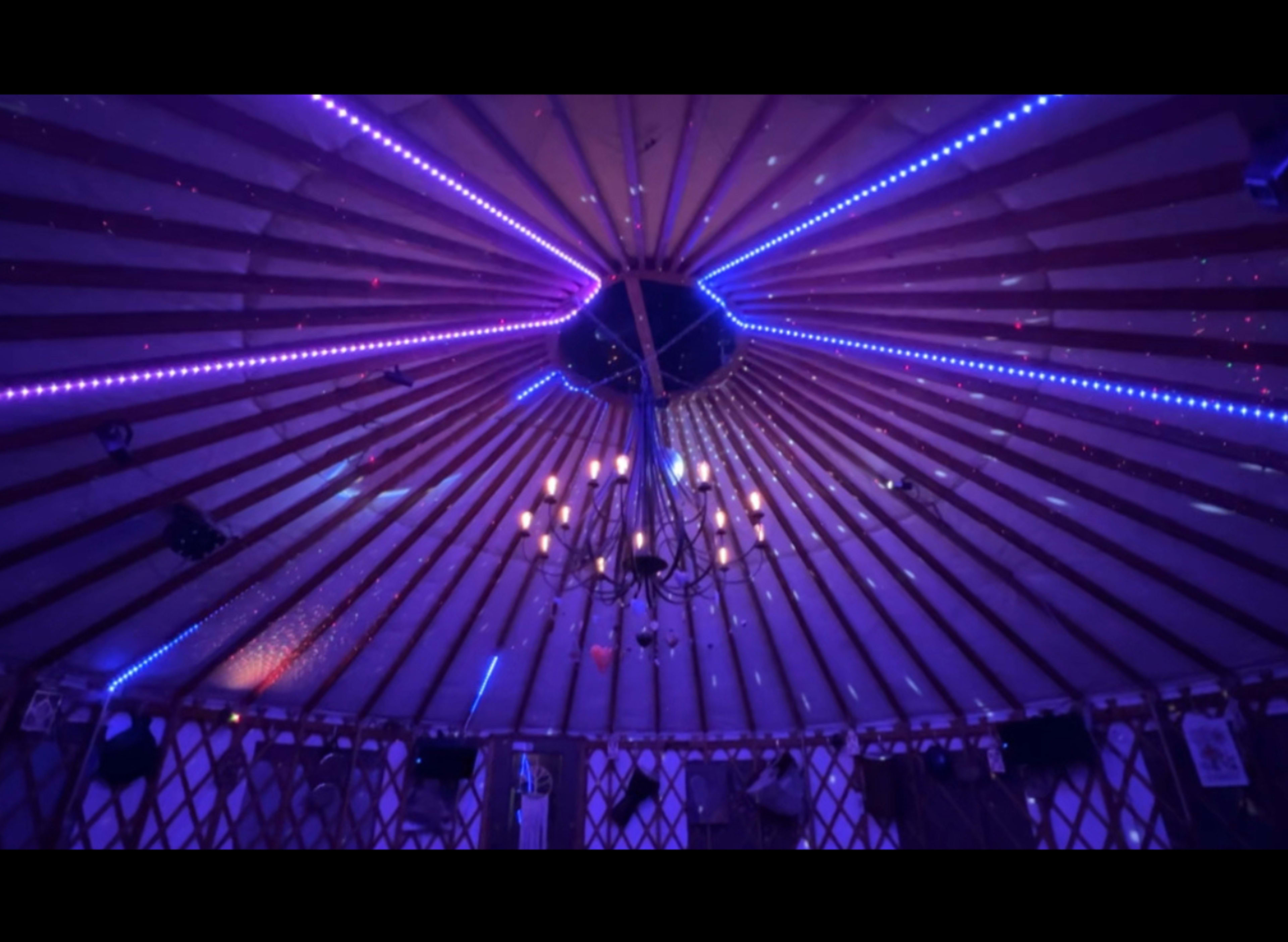 The interior of a yurt features a circular ceiling with radiating purple and blue lights, along with a central chandelier.
