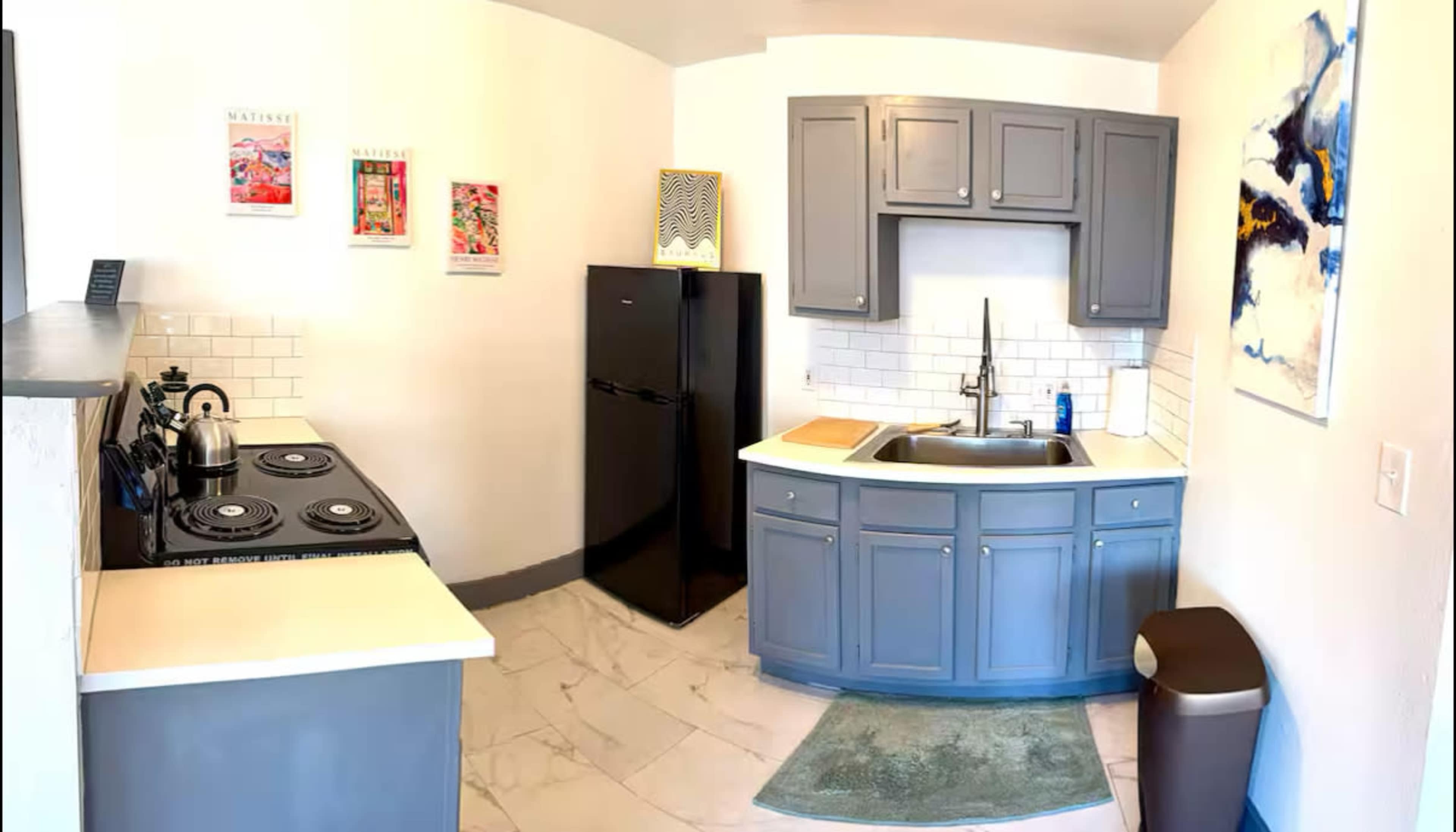 The image shows a compact kitchen featuring a black refrigerator, a sink with a gray cabinetry, a gas stove, and a small waste bin.