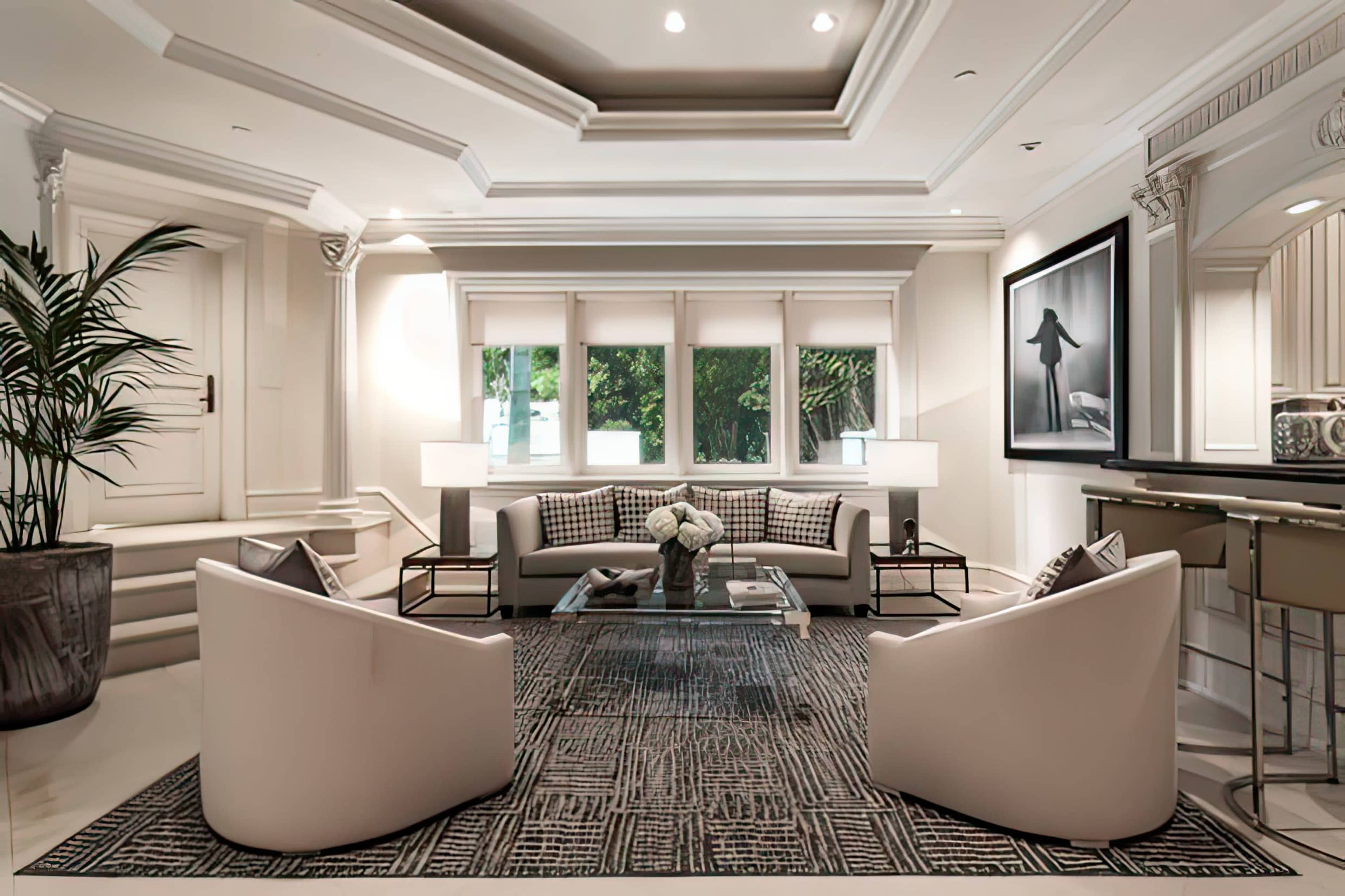 A modern living room features a gray sofa, two white chairs, a glass coffee table, and large windows with natural light.