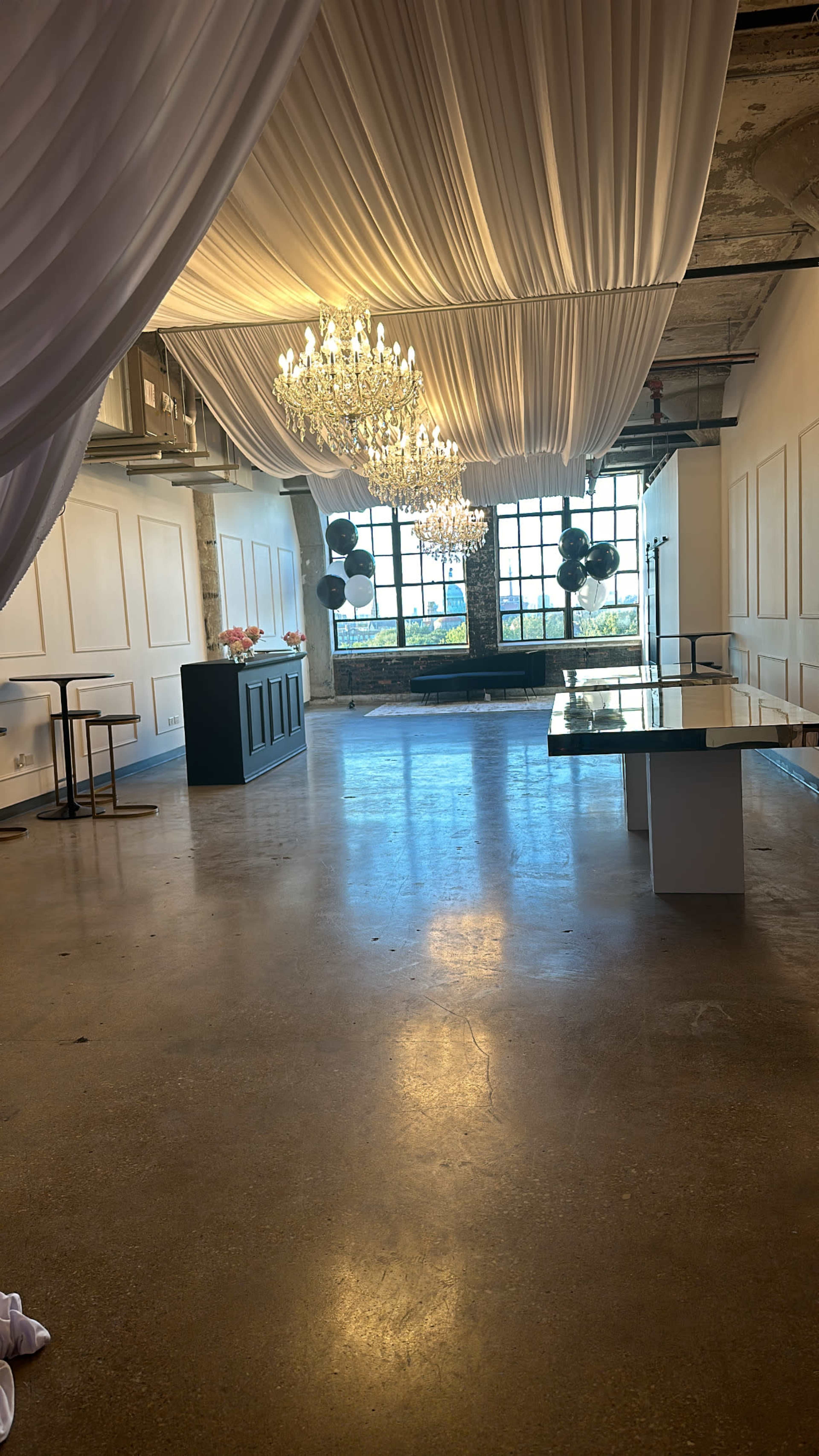 A spacious indoor venue with a polished concrete floor, featuring large chandeliers hanging from the ceiling and a decorative wall with large windows.