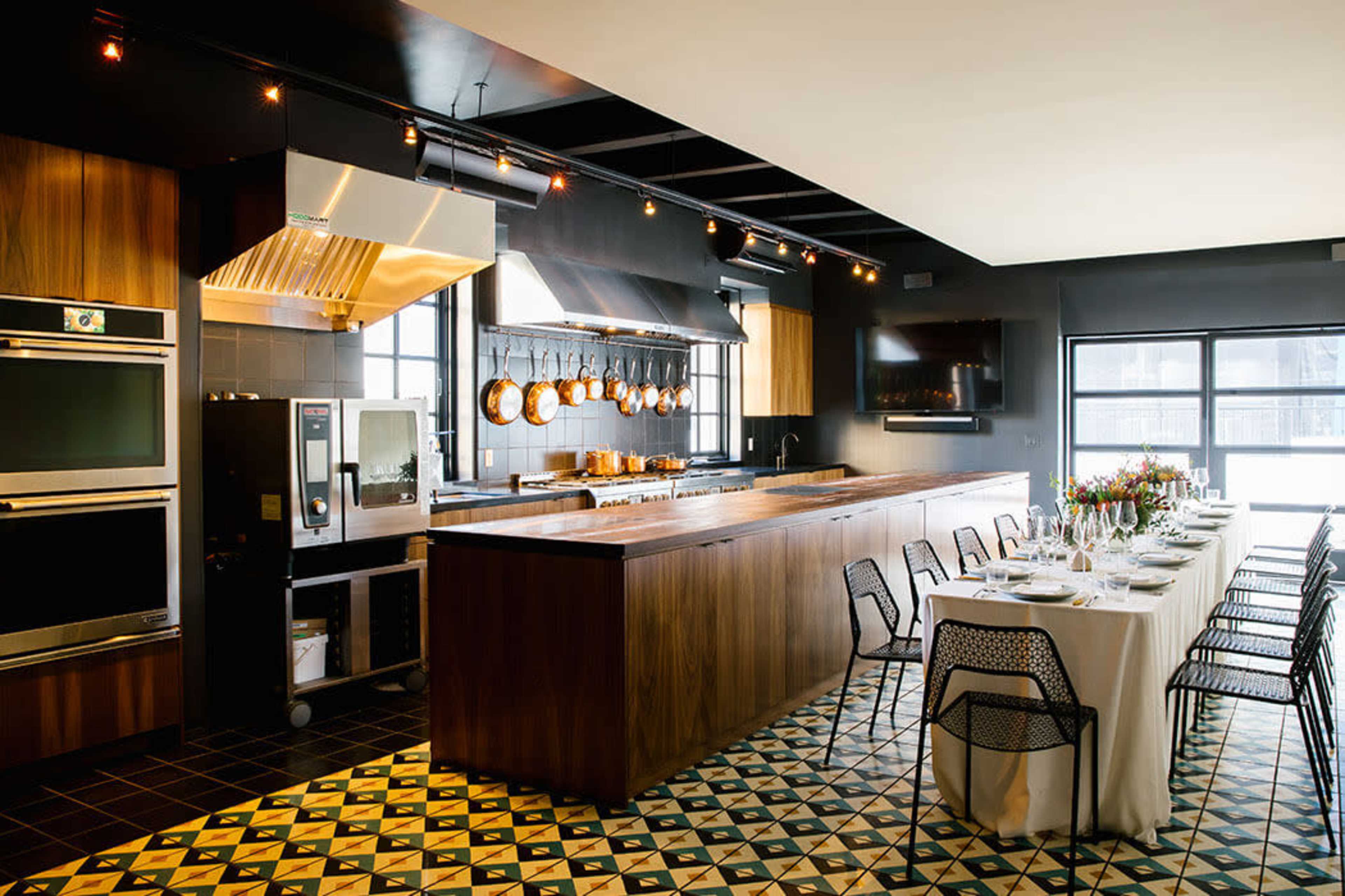 The image shows a modern kitchen with a large island, stainless steel appliances, and a dining table set for a meal.
