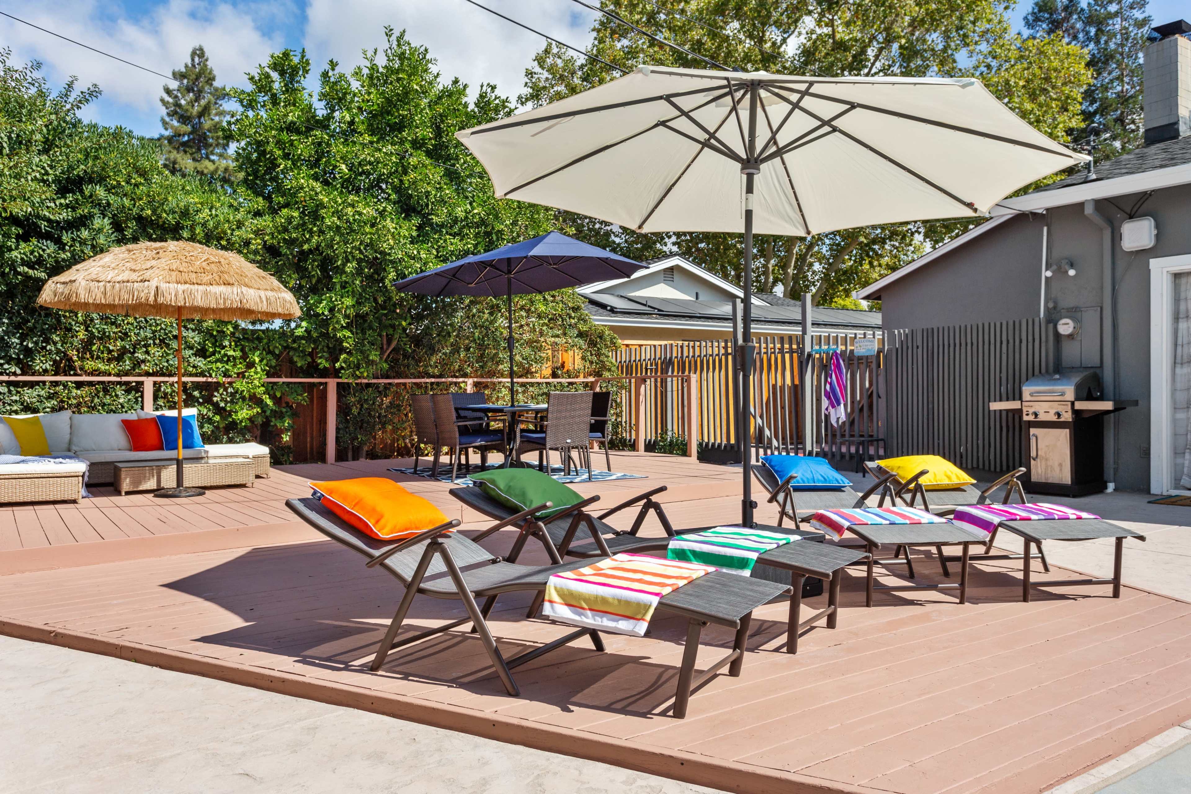 A backyard features several lounge chairs with colorful cushions, two umbrellas, a shaded seating area, and a grill on a wooden deck.