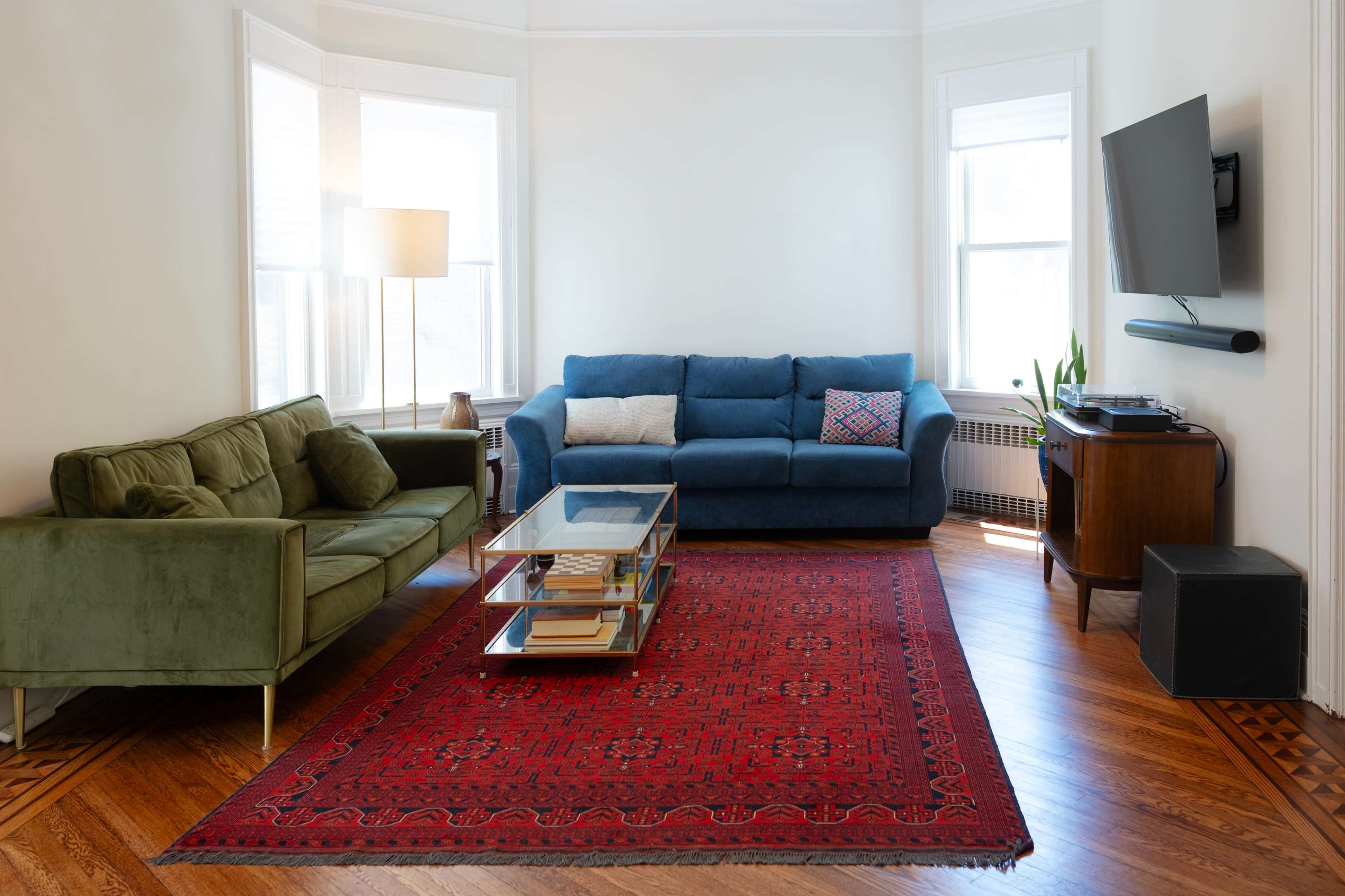 The living room features two sofas, a glass coffee table on a red area rug, and a television mounted on the wall.