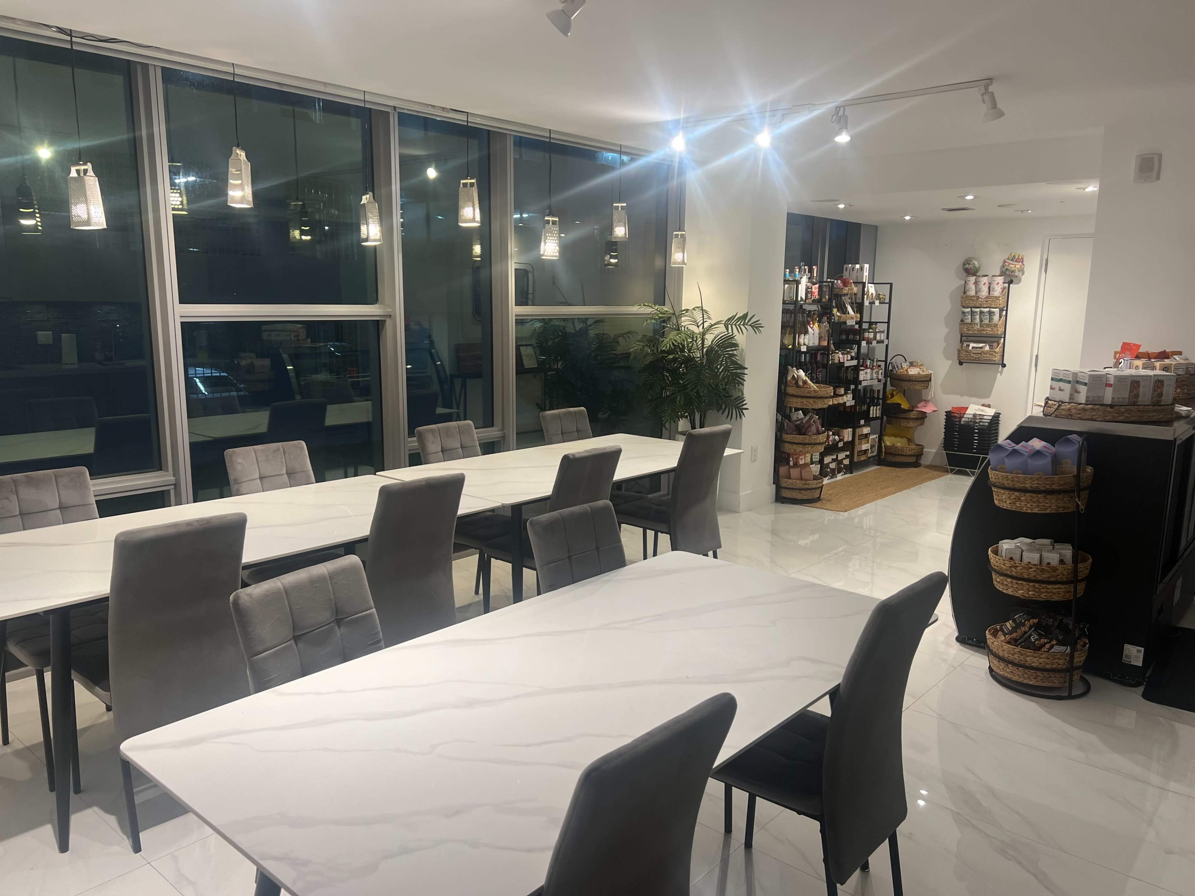 A modern dining area features several white marble tables surrounded by gray chairs, with large windows, plants, and a display shelf stocked with various products in the background.