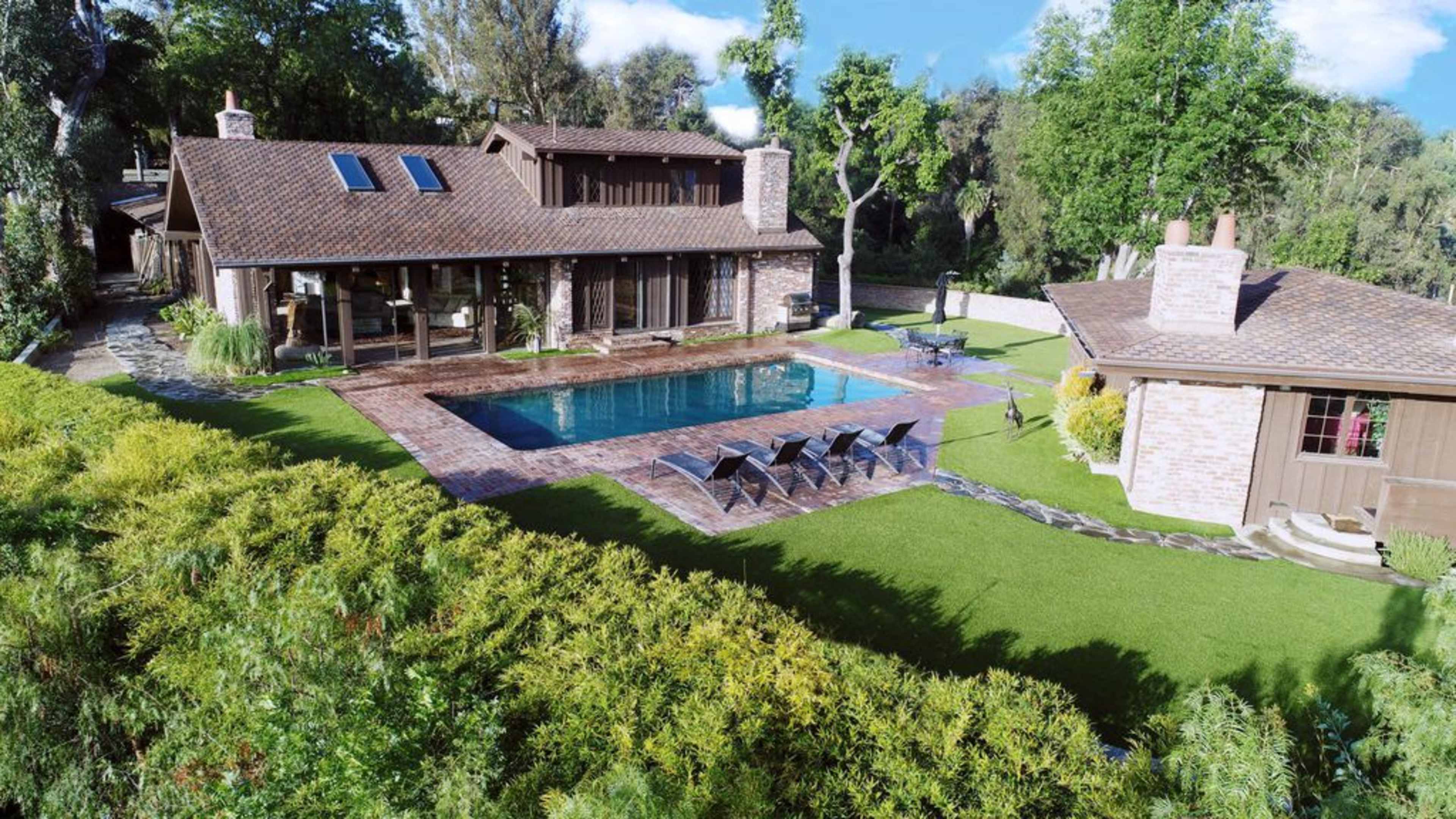 A modern house with a wooden exterior, a swimming pool surrounded by patio tiles, and lounge chairs, set in a landscaped garden with lush greenery.