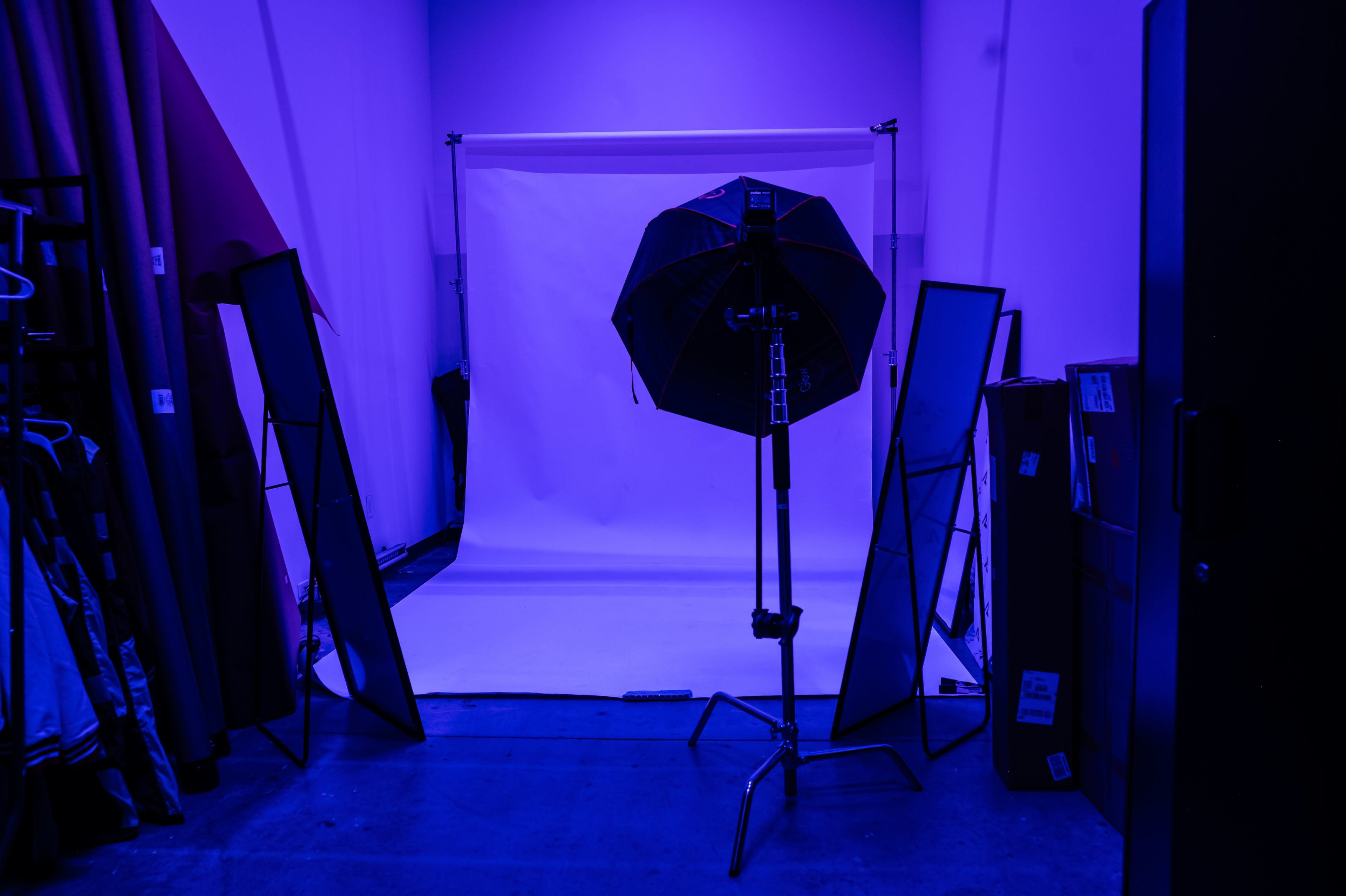 A photography studio features a white backdrop, a large softbox light, and several standing mirrors, all illuminated by blue lighting.