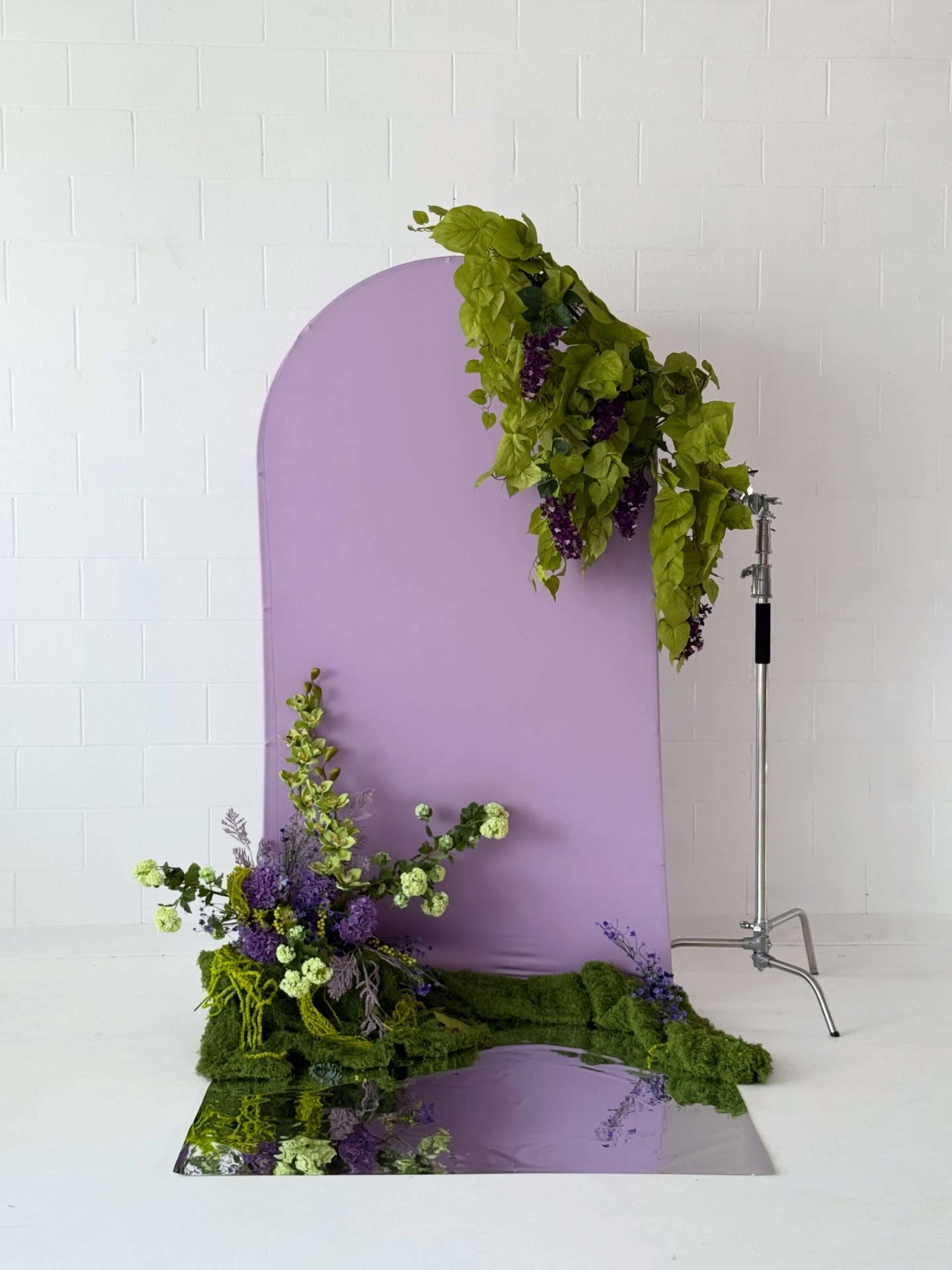 A lavender backdrop is adorned with greenery and flowers, resting on a patch of green moss and a reflective surface.