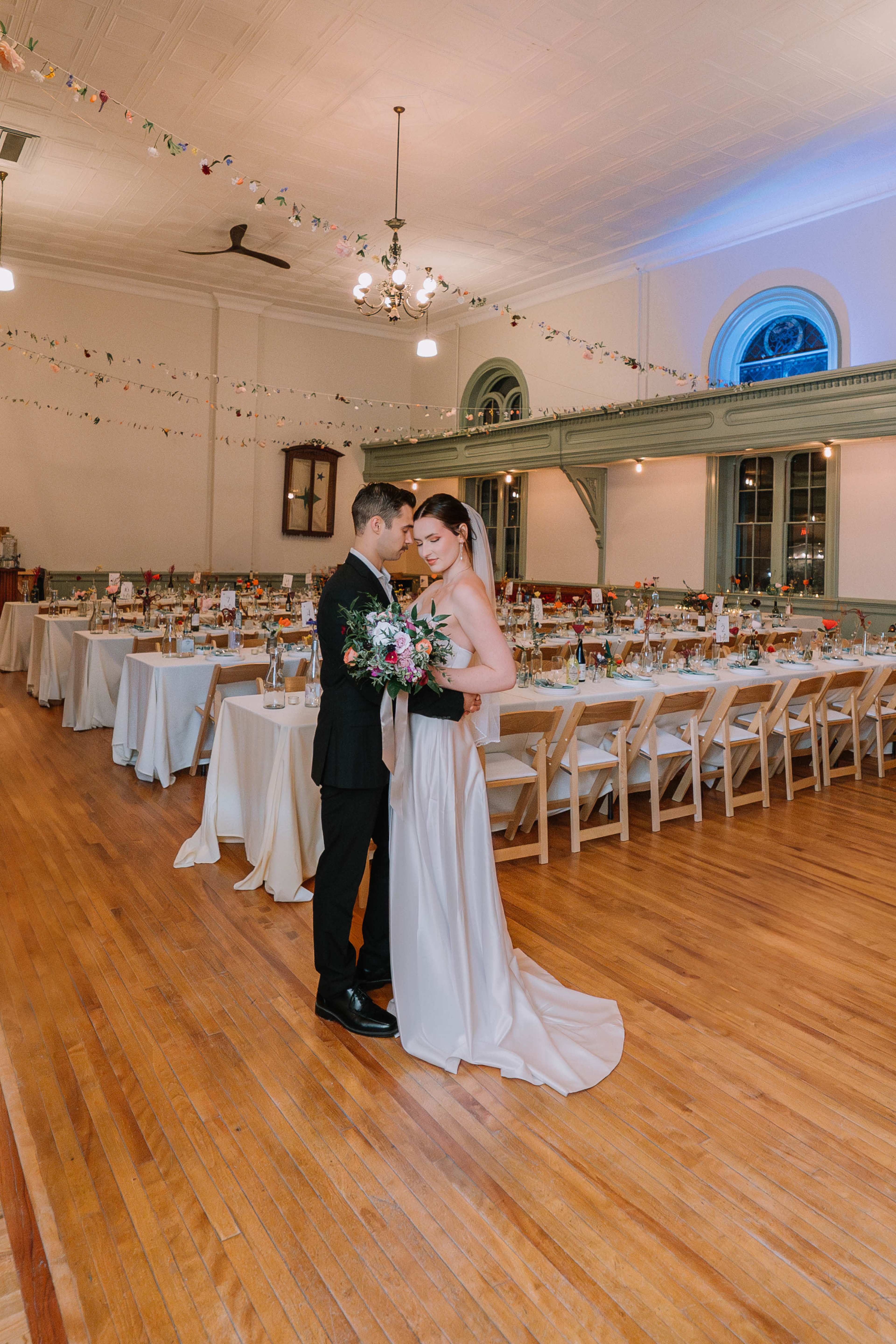 A bride and groom share a moment together in a decorated reception hall filled with tables and floral arrangements.