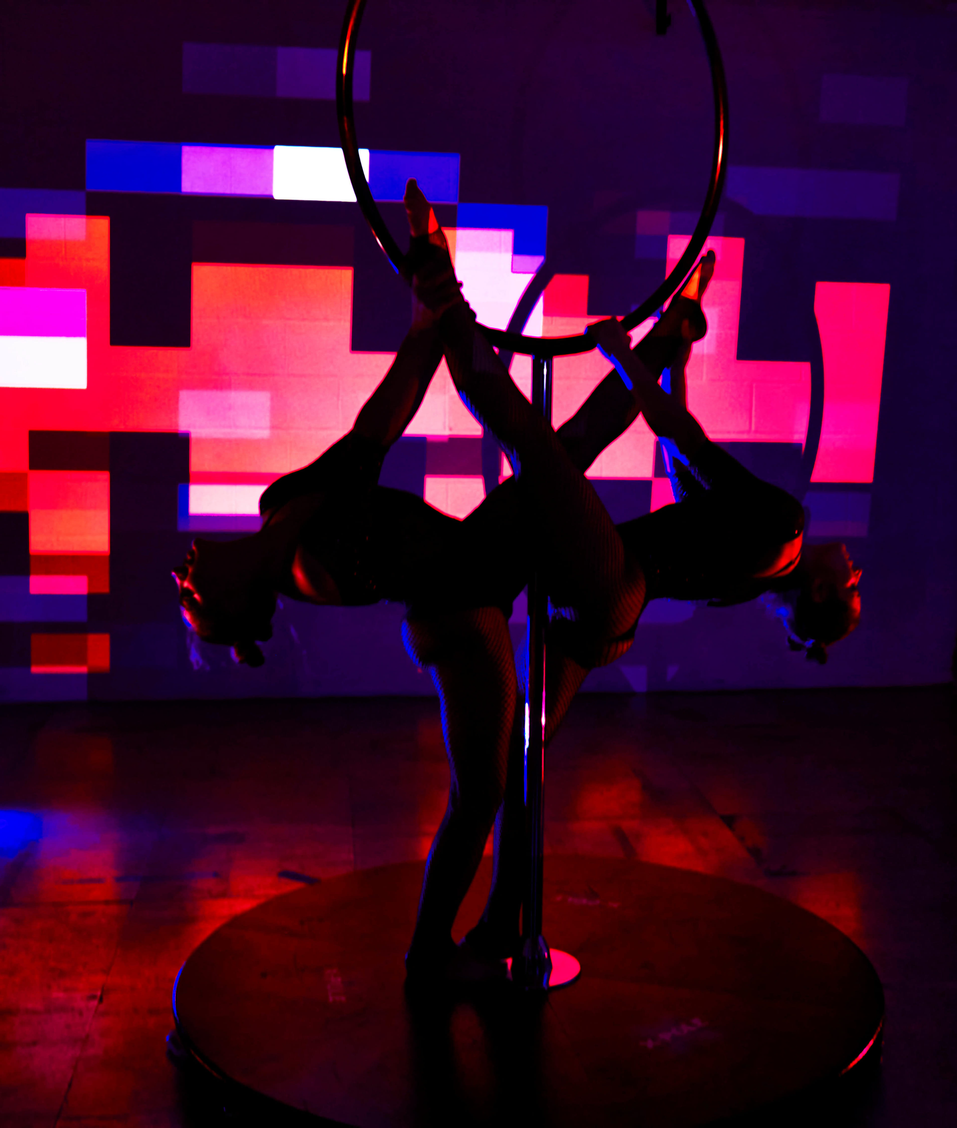 Two performers in black costumes are posing on a silver aerial hoop against a backdrop of colorful, abstract lights.