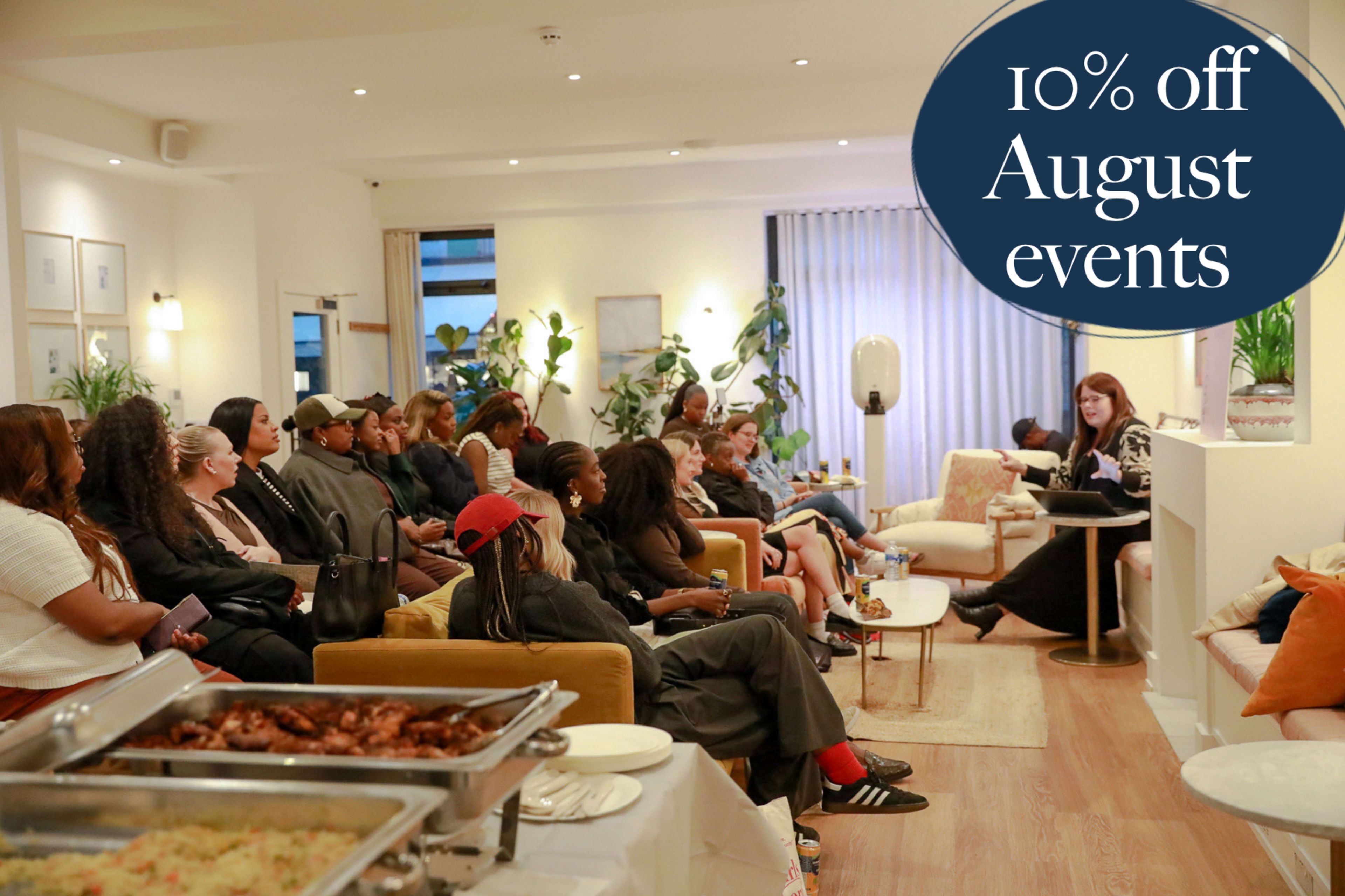 A group of people is seated in a bright, modern space, listening to a speaker, while a buffet of food is set up in the foreground.