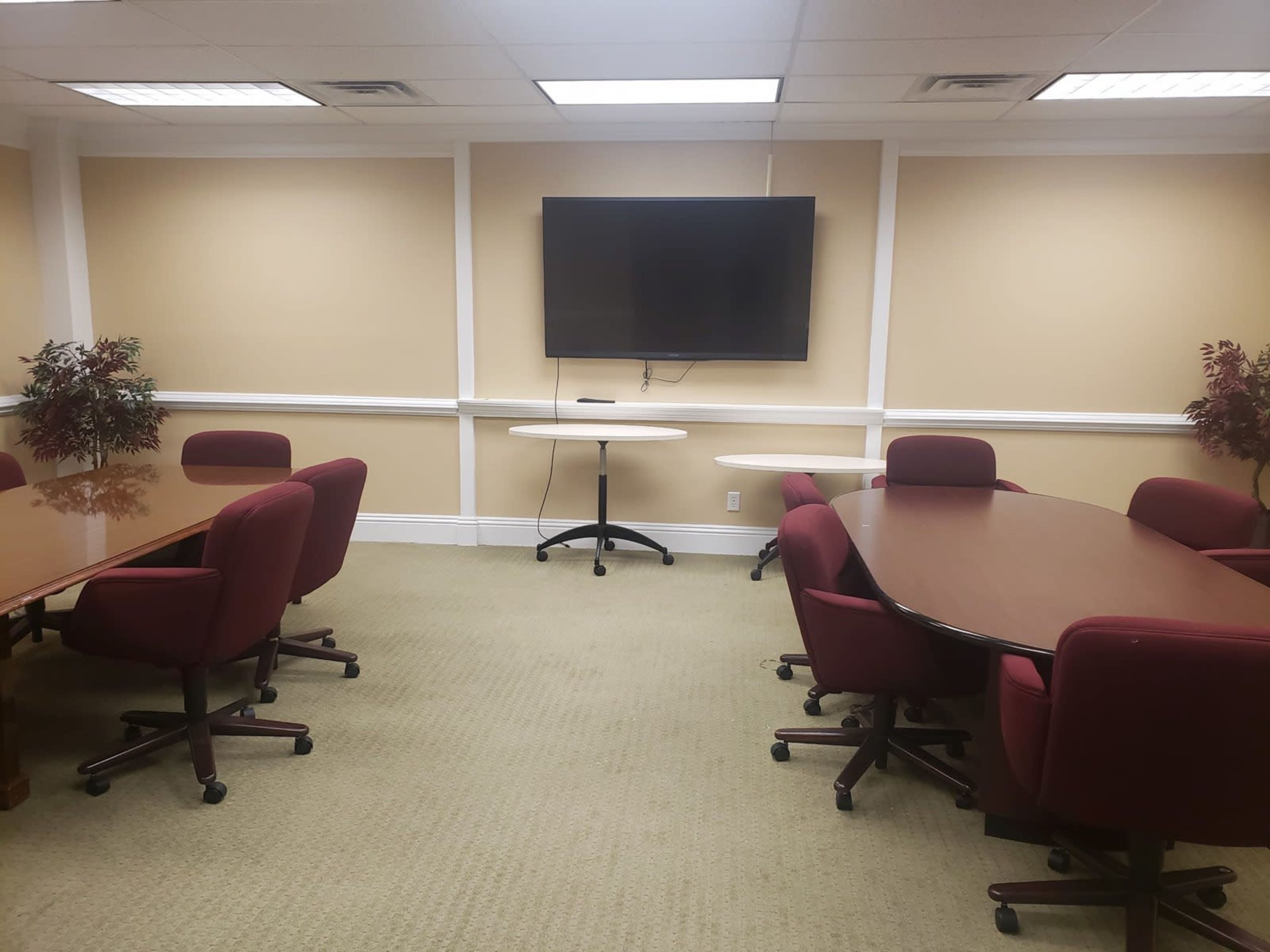 A conference room features multiple oval tables surrounded by red office chairs, with a television mounted on the wall.