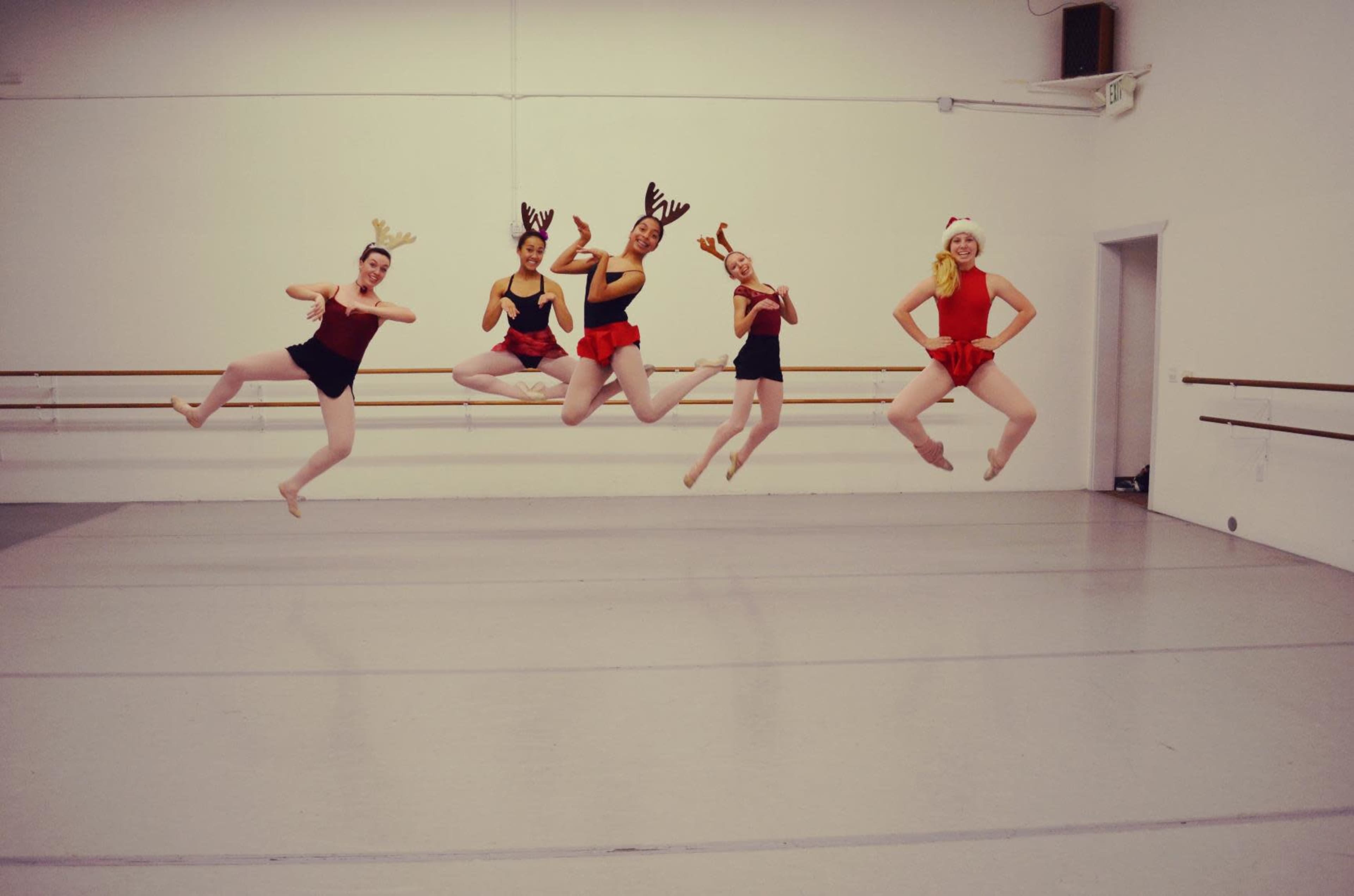 Five dancers in festive attire jump in mid-air inside a dance studio.
