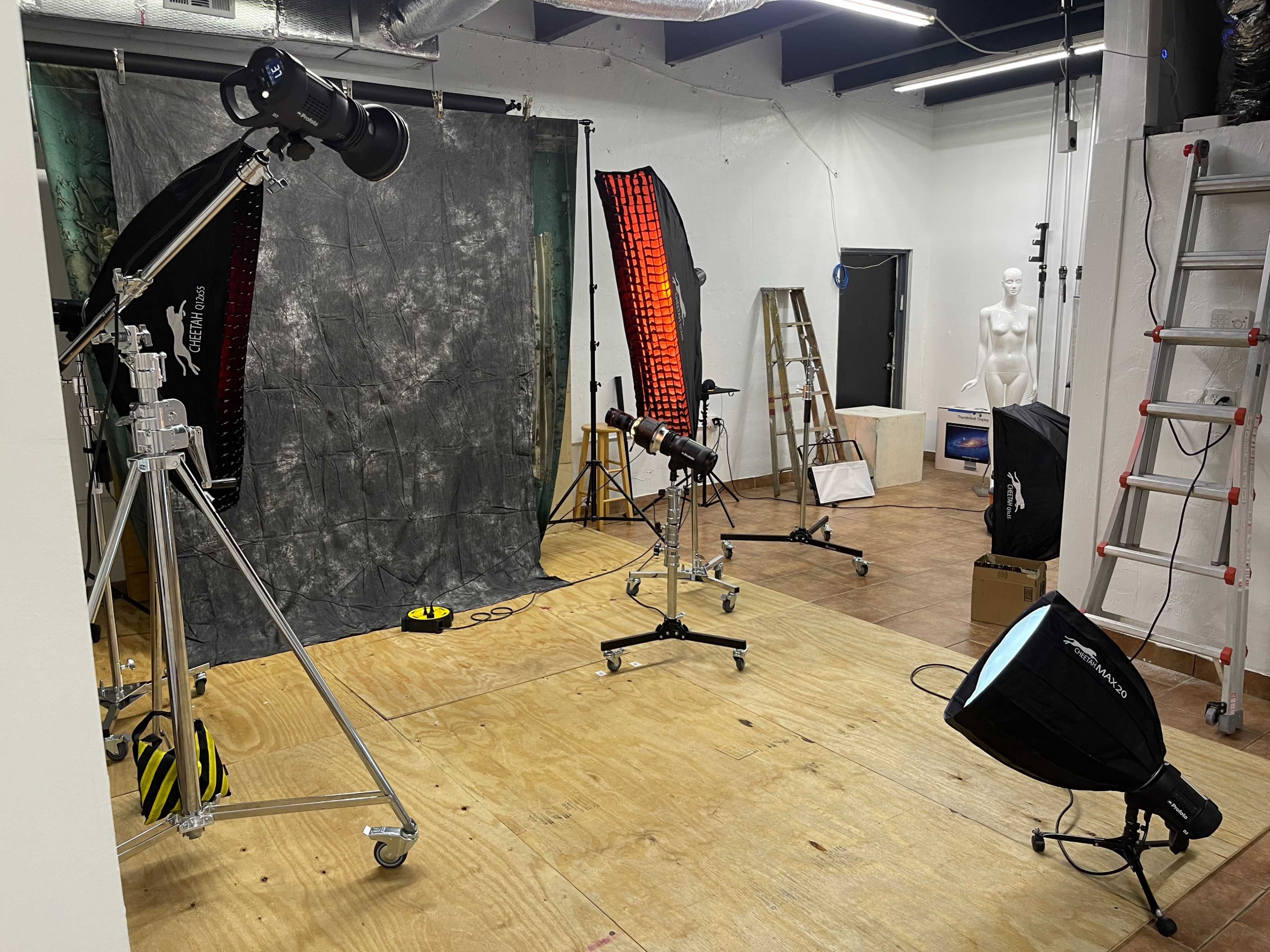 The image shows a photography studio equipped with various lighting equipment, a backdrop, a mannequin, and ladders.
