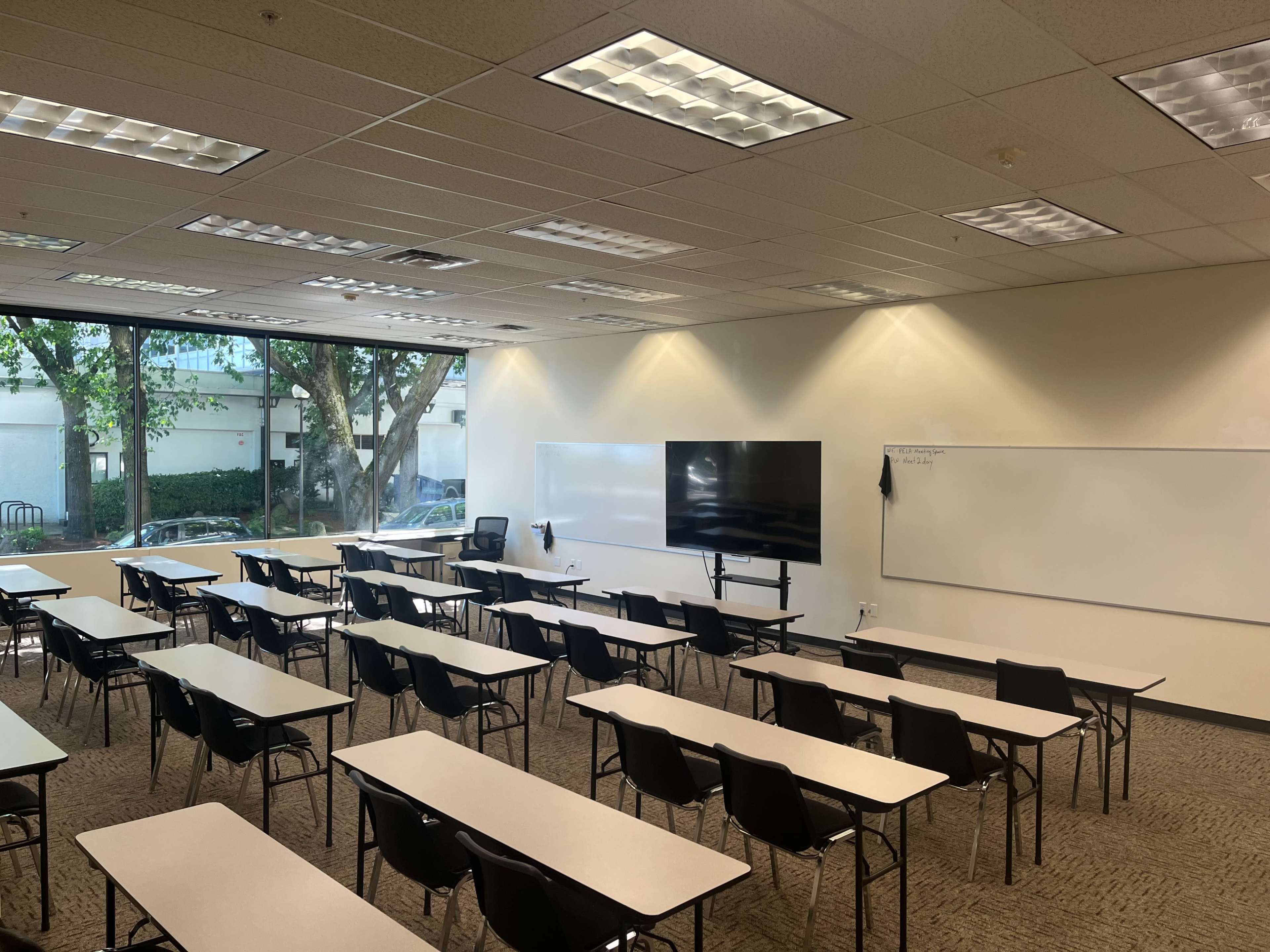 The image shows a neatly arranged classroom with multiple rows of desks facing a large screen and whiteboards, surrounded by windows letting in natural light.