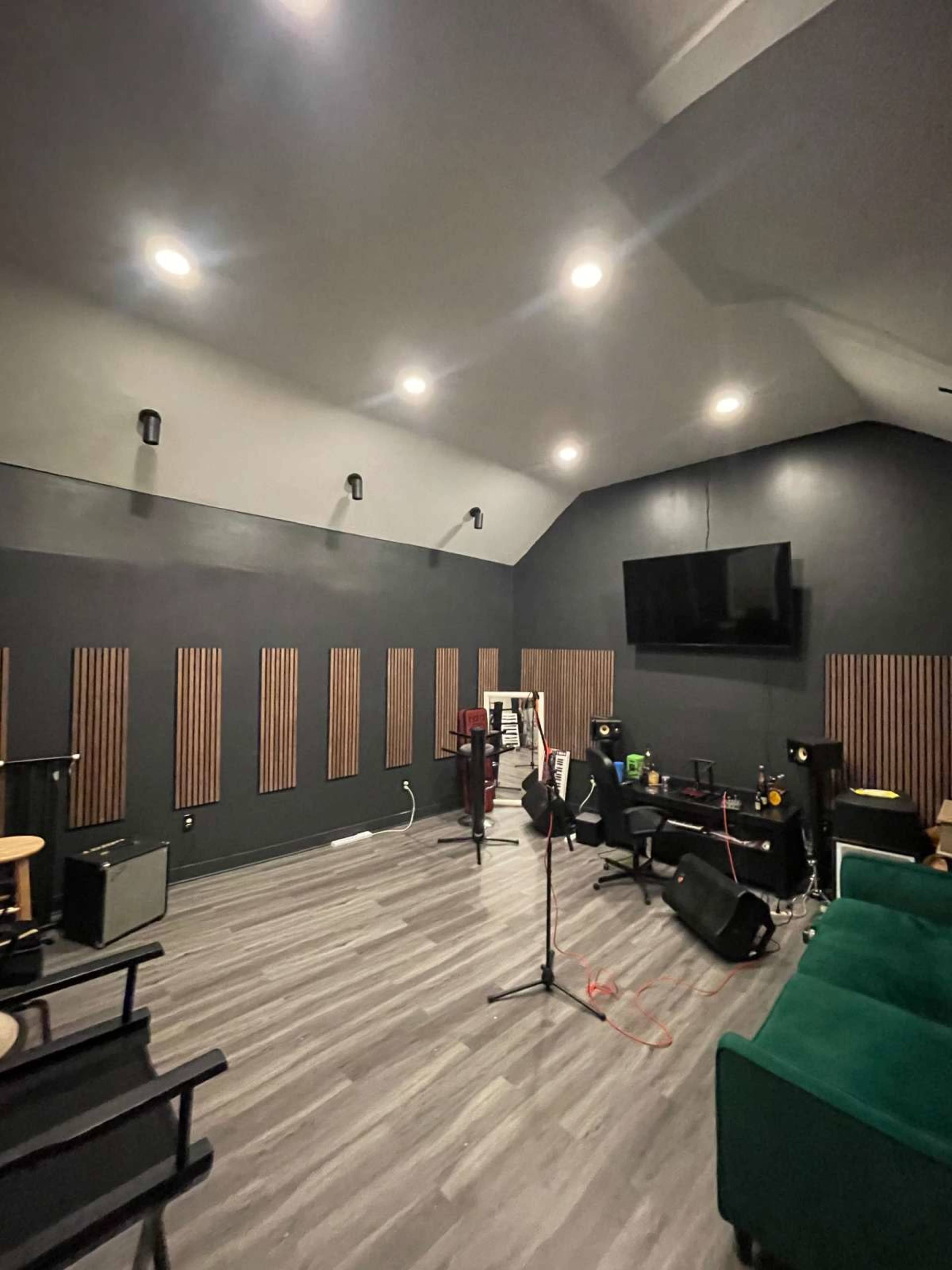 The image shows a music rehearsal studio featuring a microphone stand, seating, and sound-absorbing wall panels, with a television mounted on the wall.