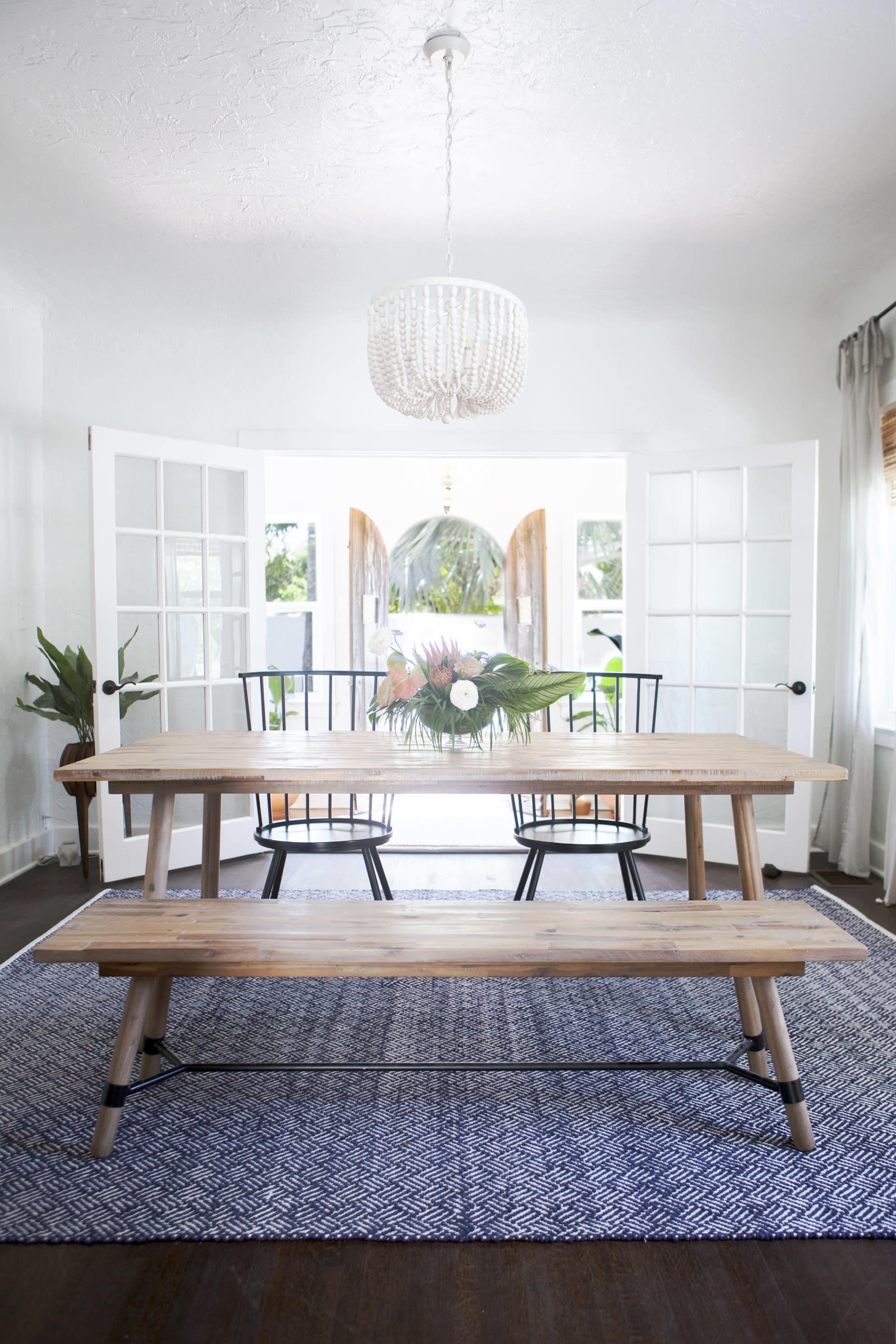 A wooden dining table with a floral centerpiece and black chairs is set in a bright room with open French doors leading to an outdoor view.