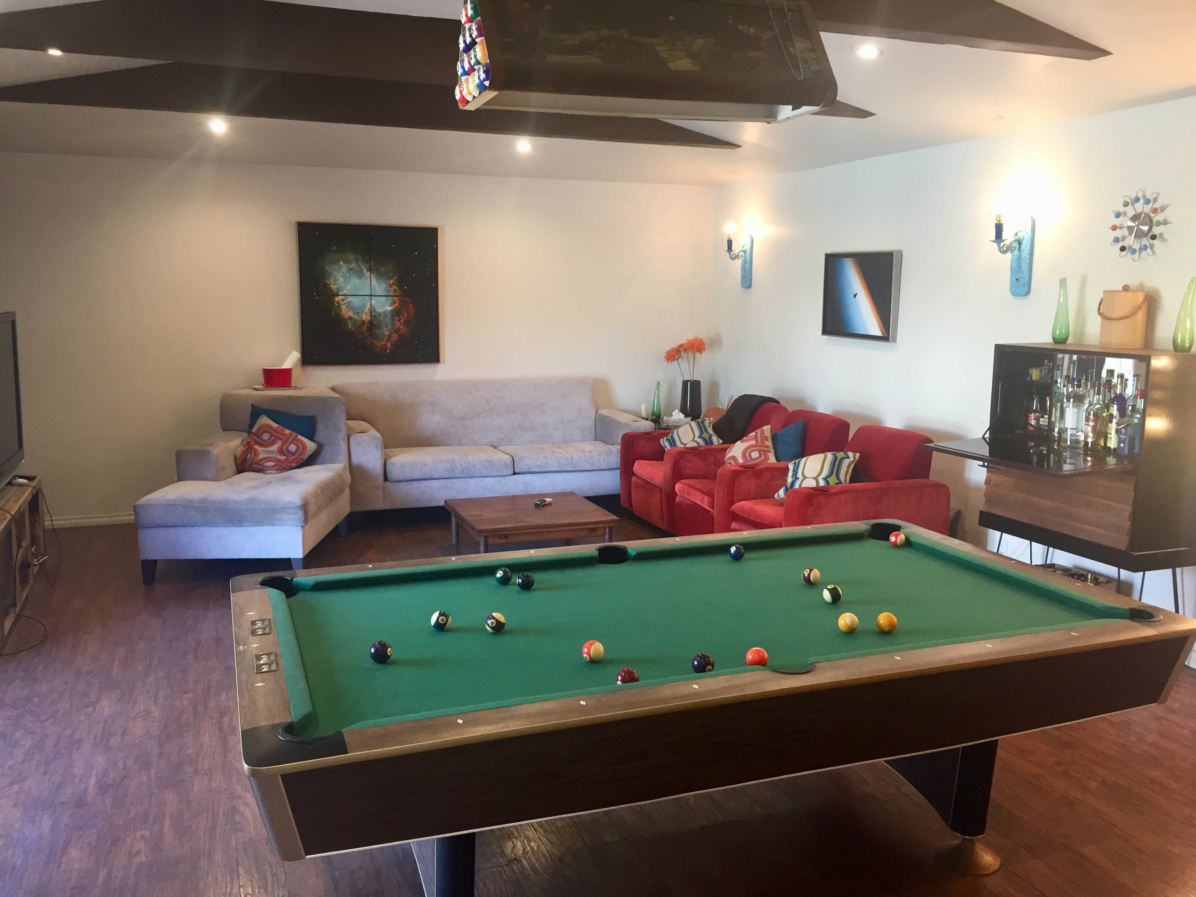 The image shows a game room featuring a pool table, a sofa, two red chairs, and a bar cabinet against the wall.