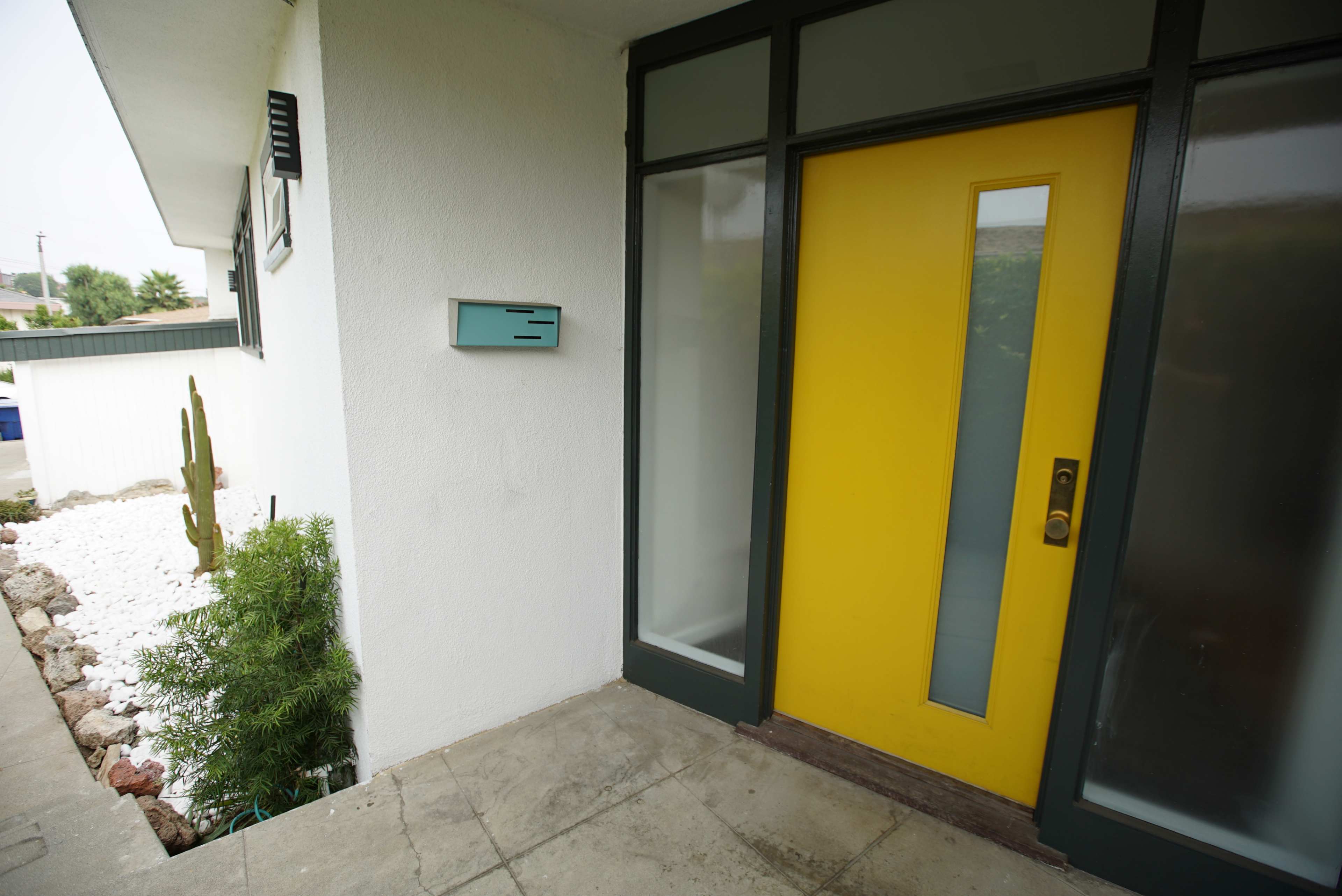 The image shows an entryway featuring a bright yellow door next to a light blue mailbox, with landscaping elements including white gravel and a cactus.