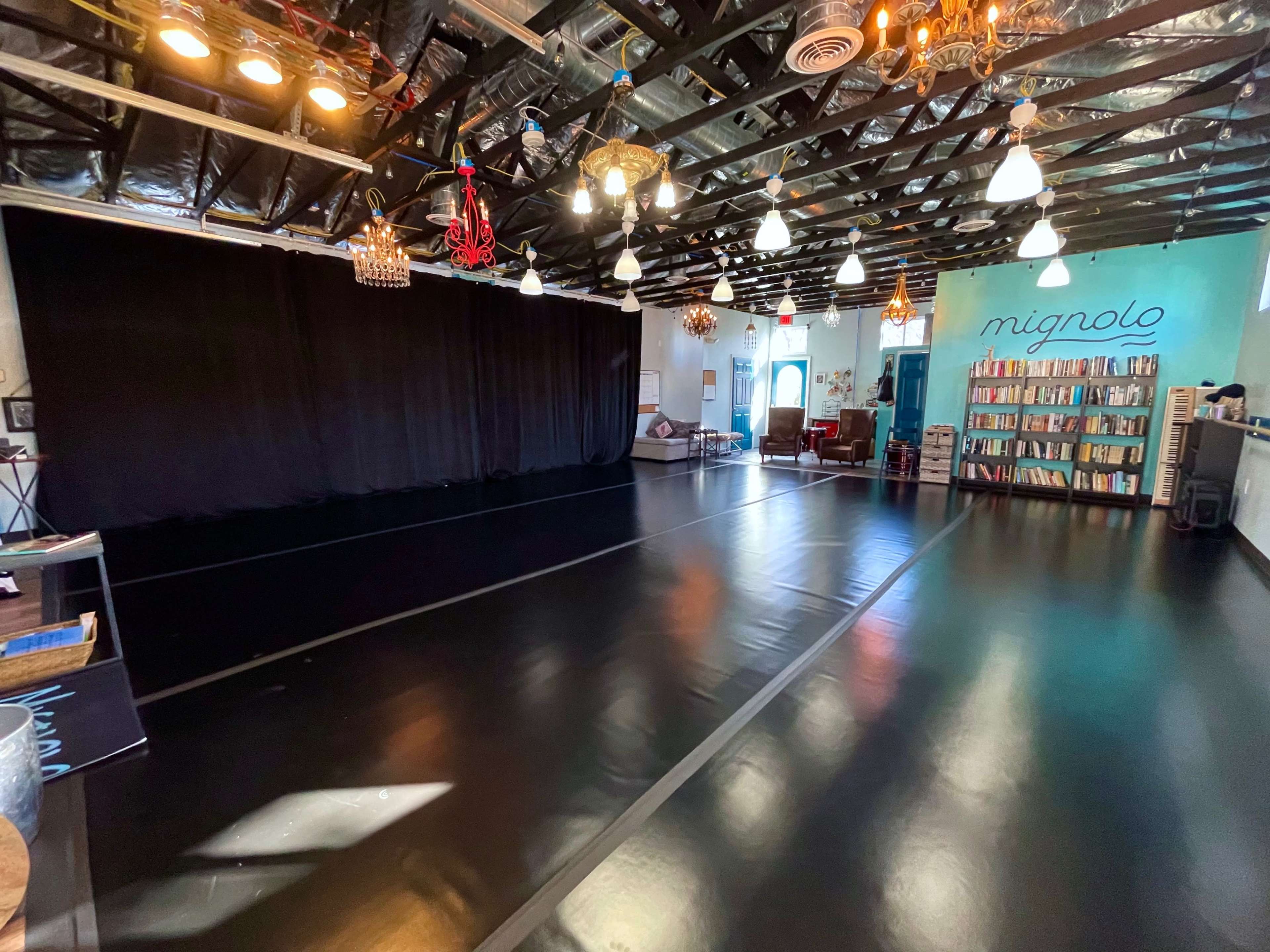 A spacious studio with black flooring, a large black curtain on one side, and a bookshelf filled with books against a wall on the other side, illuminated by various hanging lights.