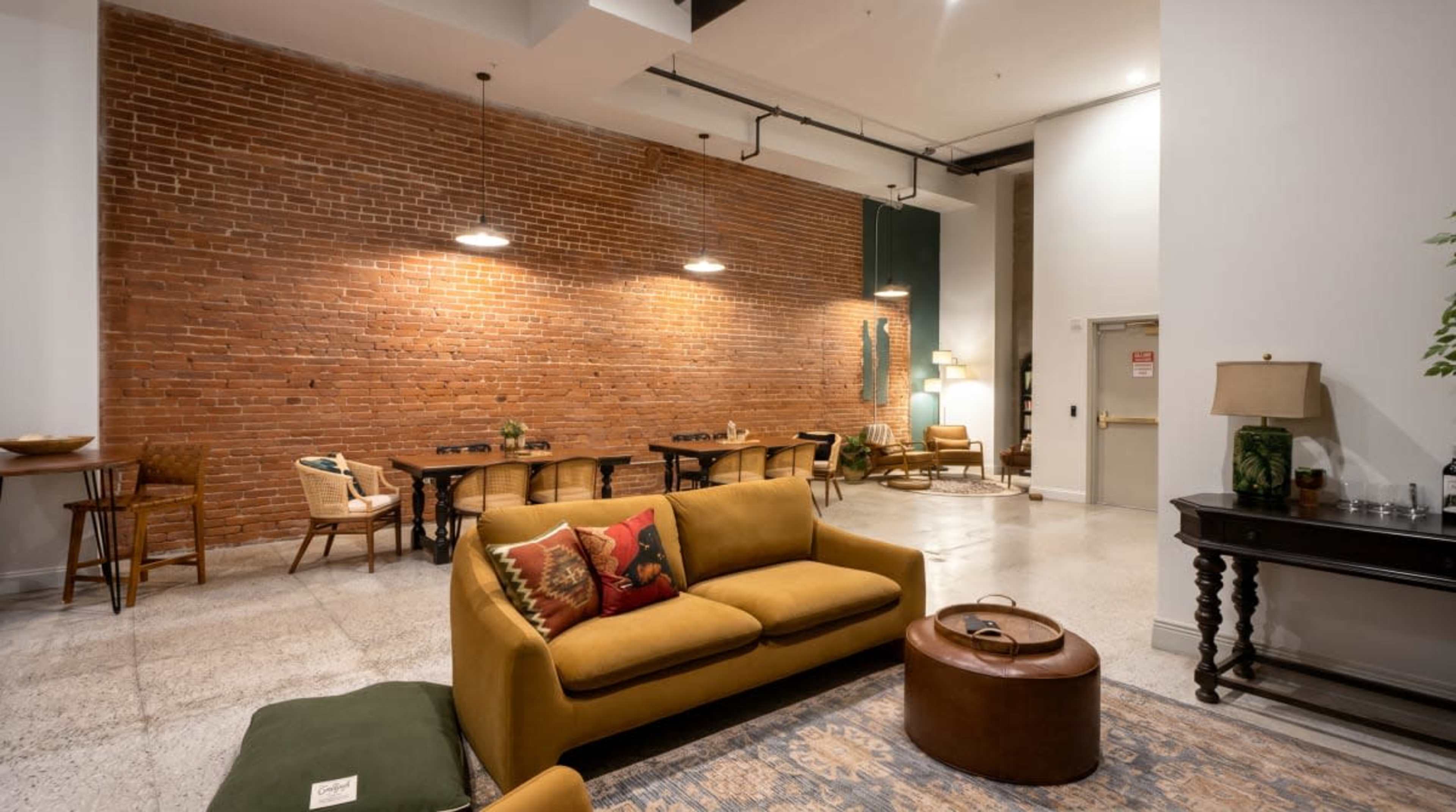 The scene features a stylish, modern living space with exposed brick walls, a seating area with a sofa and round coffee table, and a dining area with wooden chairs and a long table.