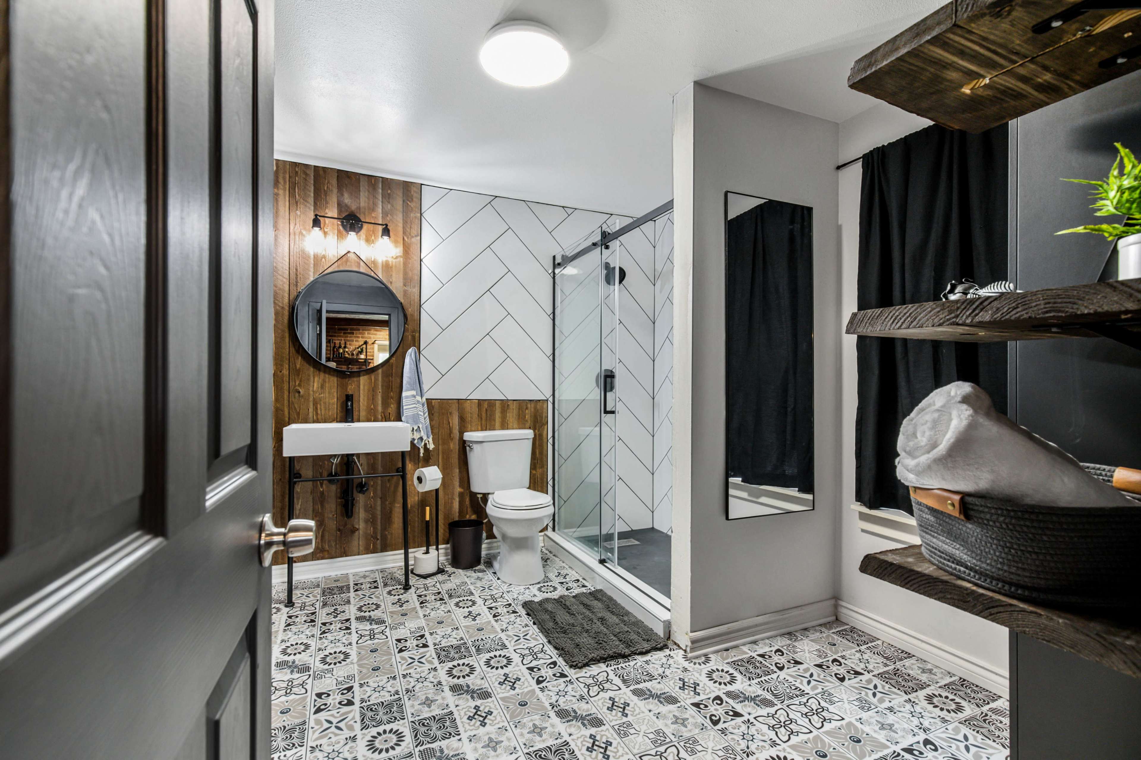 A modern bathroom featuring a glass shower, a white toilet, a sink with a round mirror, and patterned floor tiles.