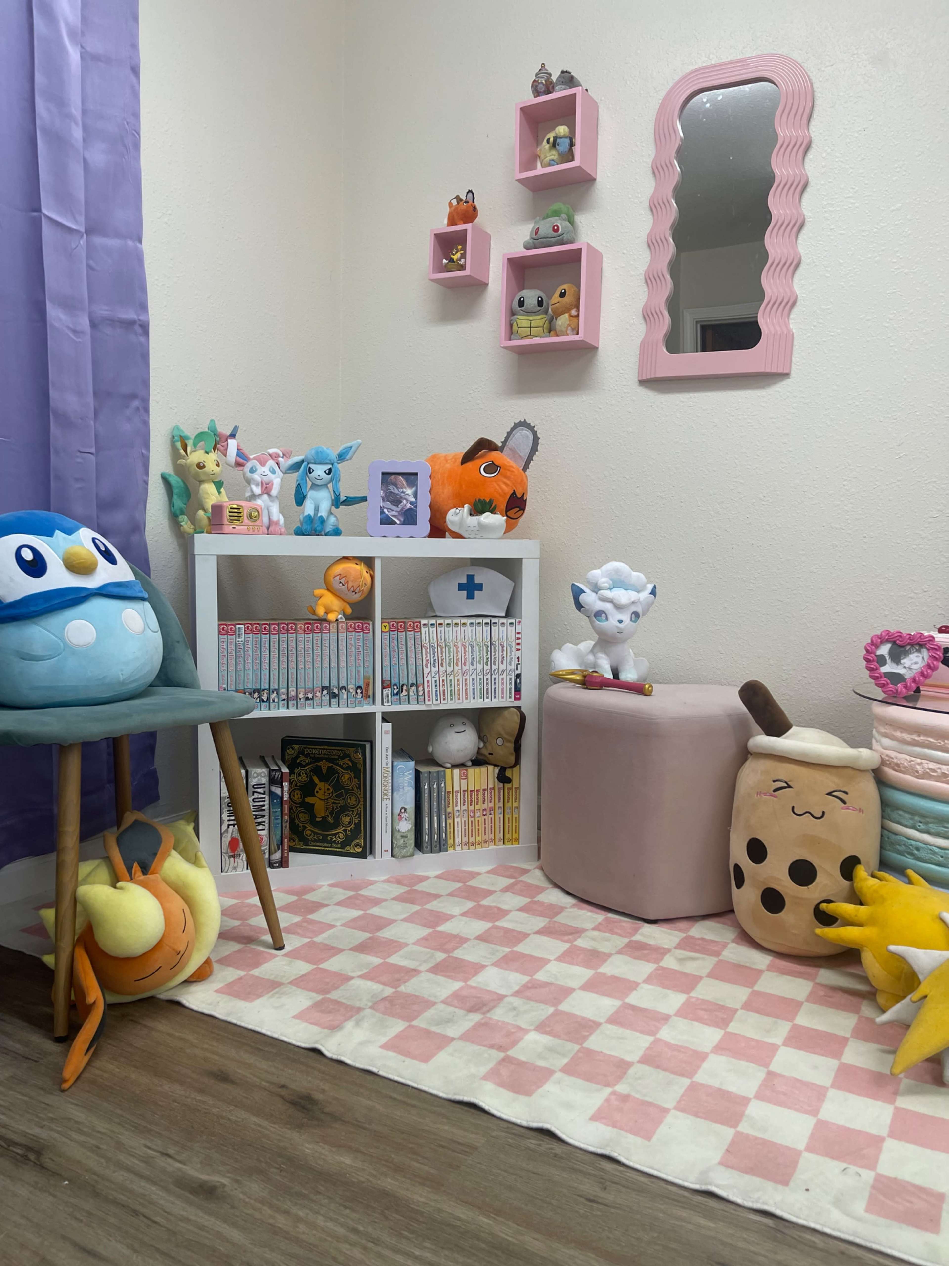 The scene features a colorful corner of a room with plush toys, manga on a white bookshelf, a round pink stool, and a mirror with a wavy frame.