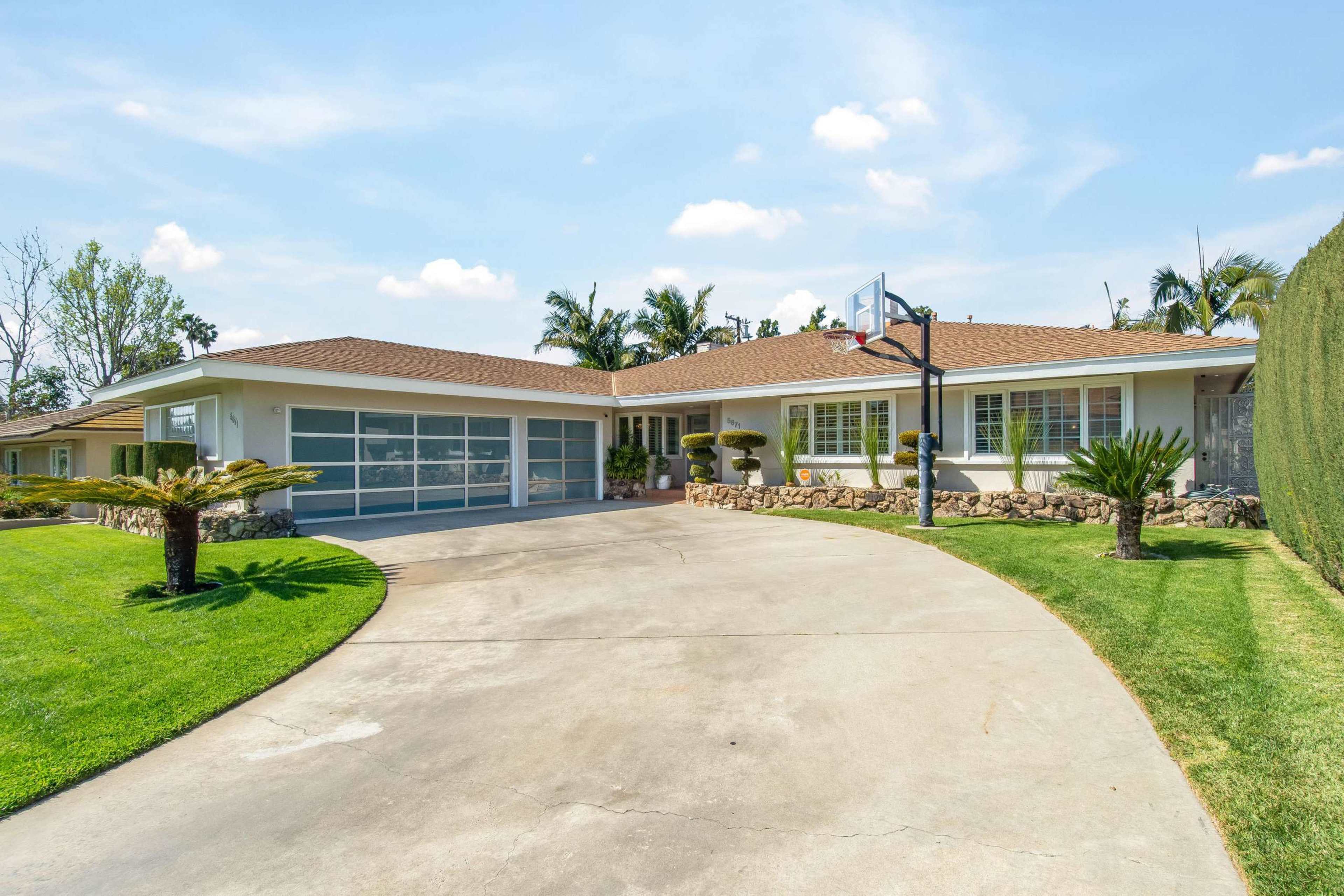 A single-story house with a basketball hoop in the driveway, surrounded by neatly manicured lawns and palm trees.