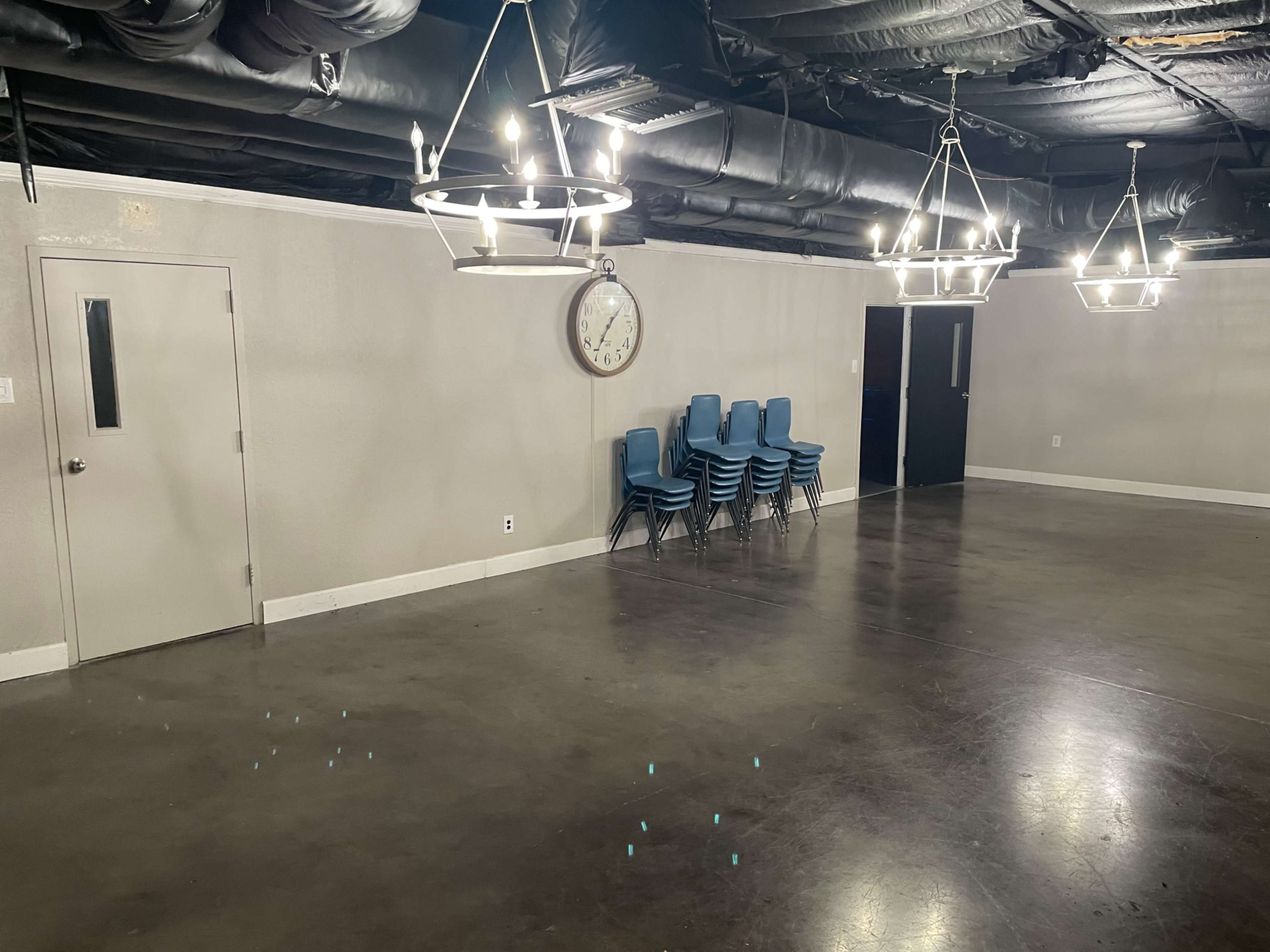 The image shows an empty room with a polished floor, six hanging light fixtures, a large clock on the wall, and a stack of blue chairs in one corner.