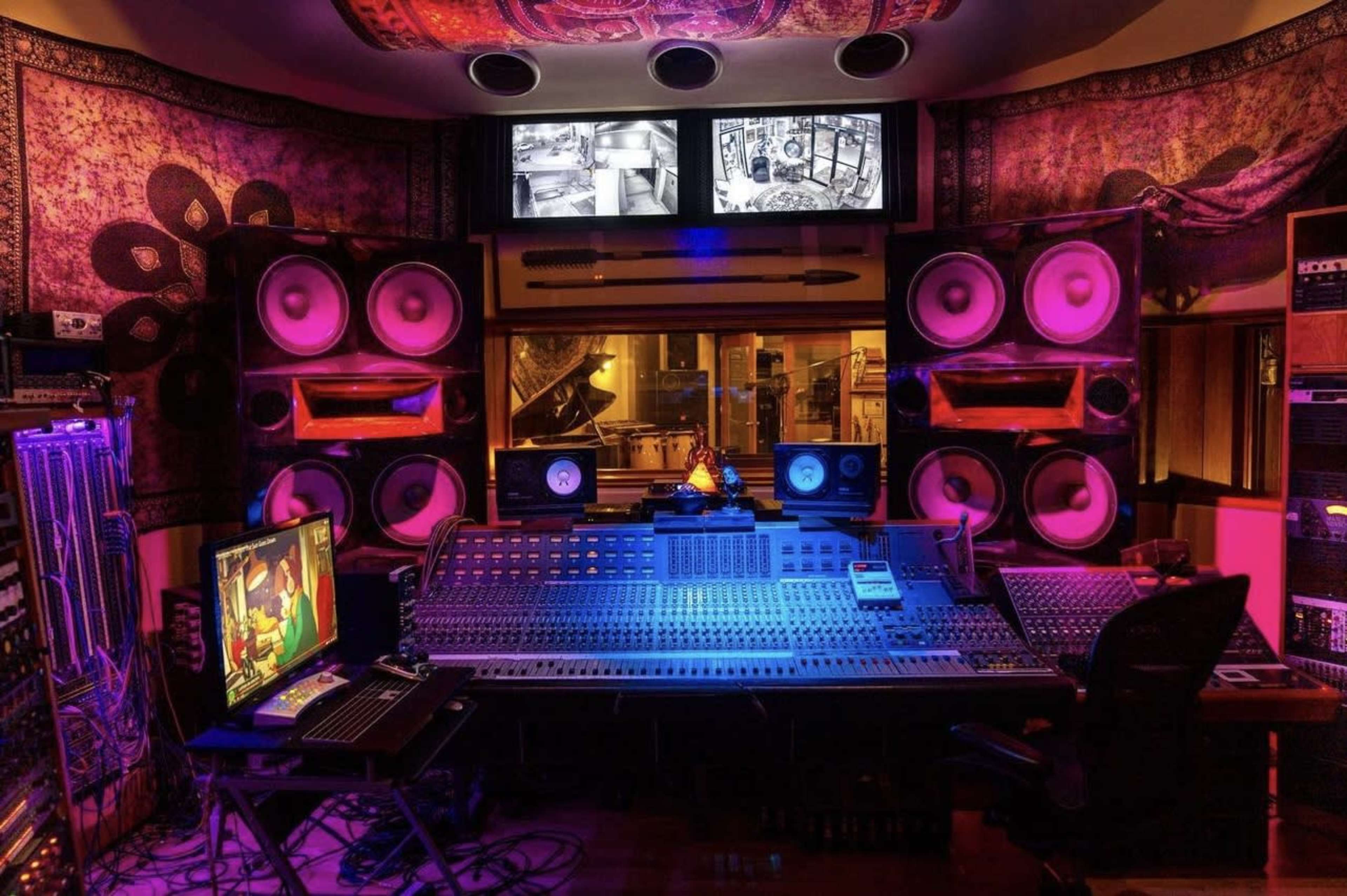 A music recording studio featuring a large mixing console surrounded by multiple speakers, colorful lighting, and monitors displaying various visuals.