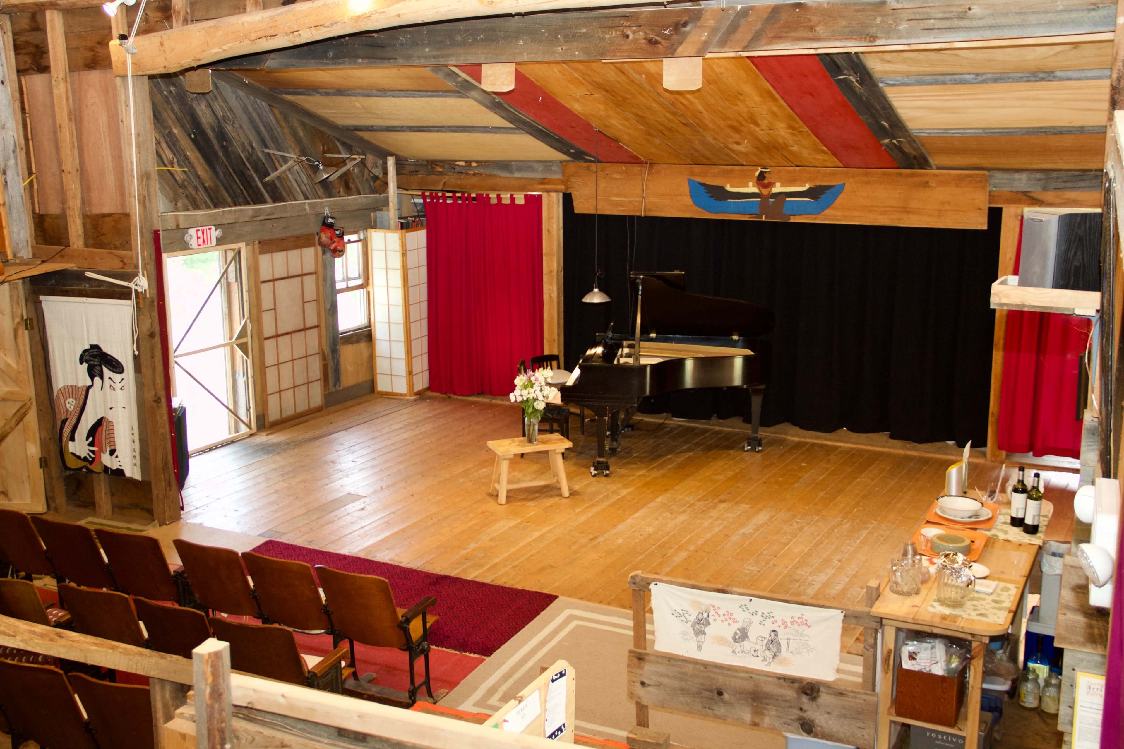The image shows a wooden performance venue featuring a grand piano on stage, surrounded by red curtains, with rows of seated audience chairs in the foreground.