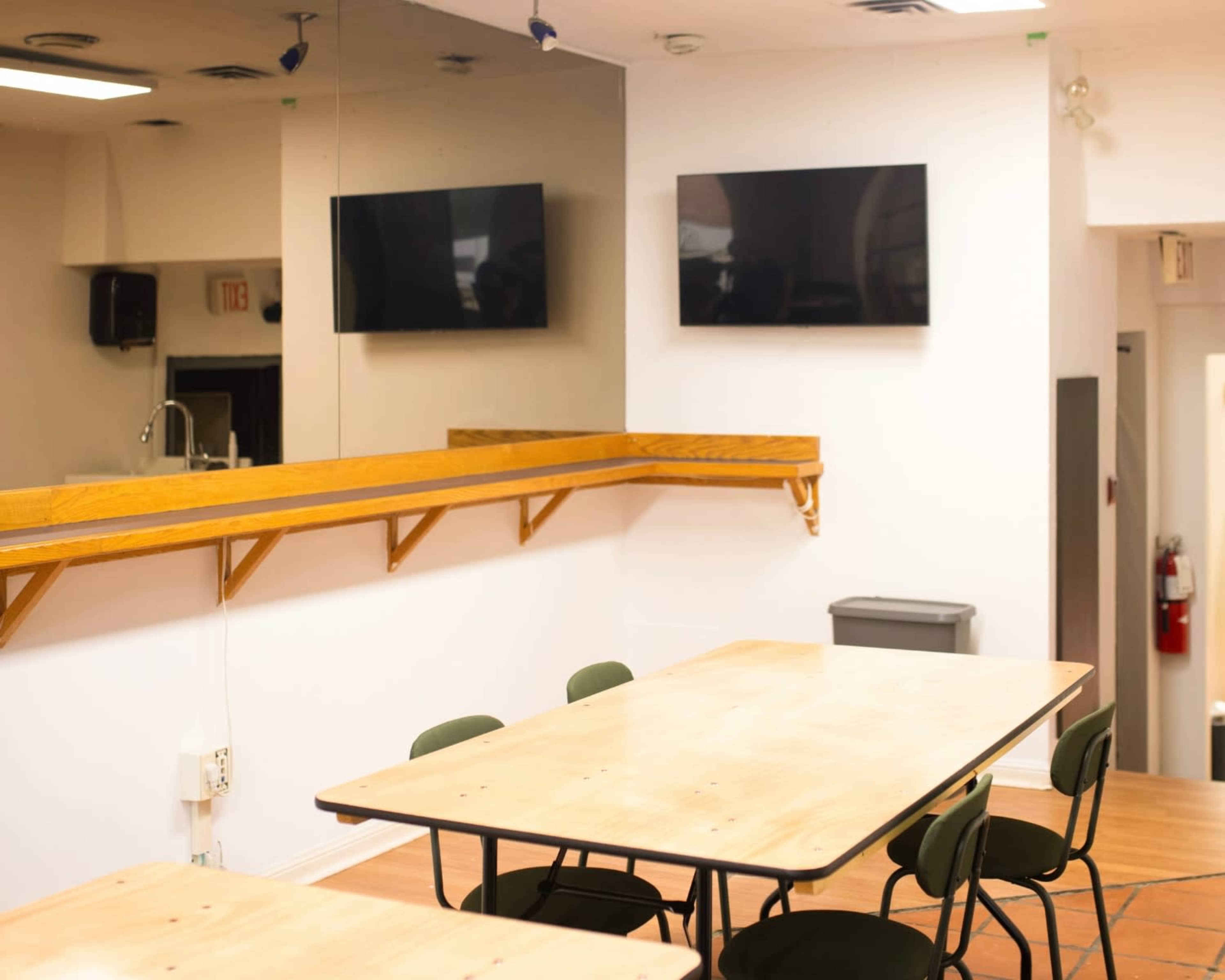 The image shows a simple room with two television screens mounted on the wall, a long wooden shelf, and several tables with chairs arranged below.