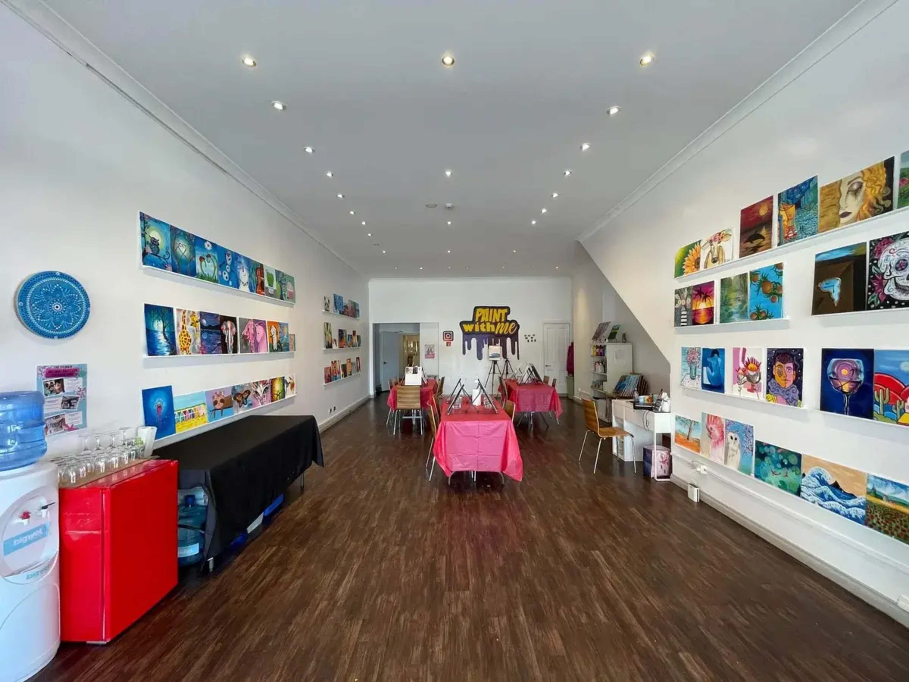 The image depicts a brightly lit art studio featuring tables with pink tablecloths, numerous framed artworks on white walls, and a refreshment area with a water cooler.