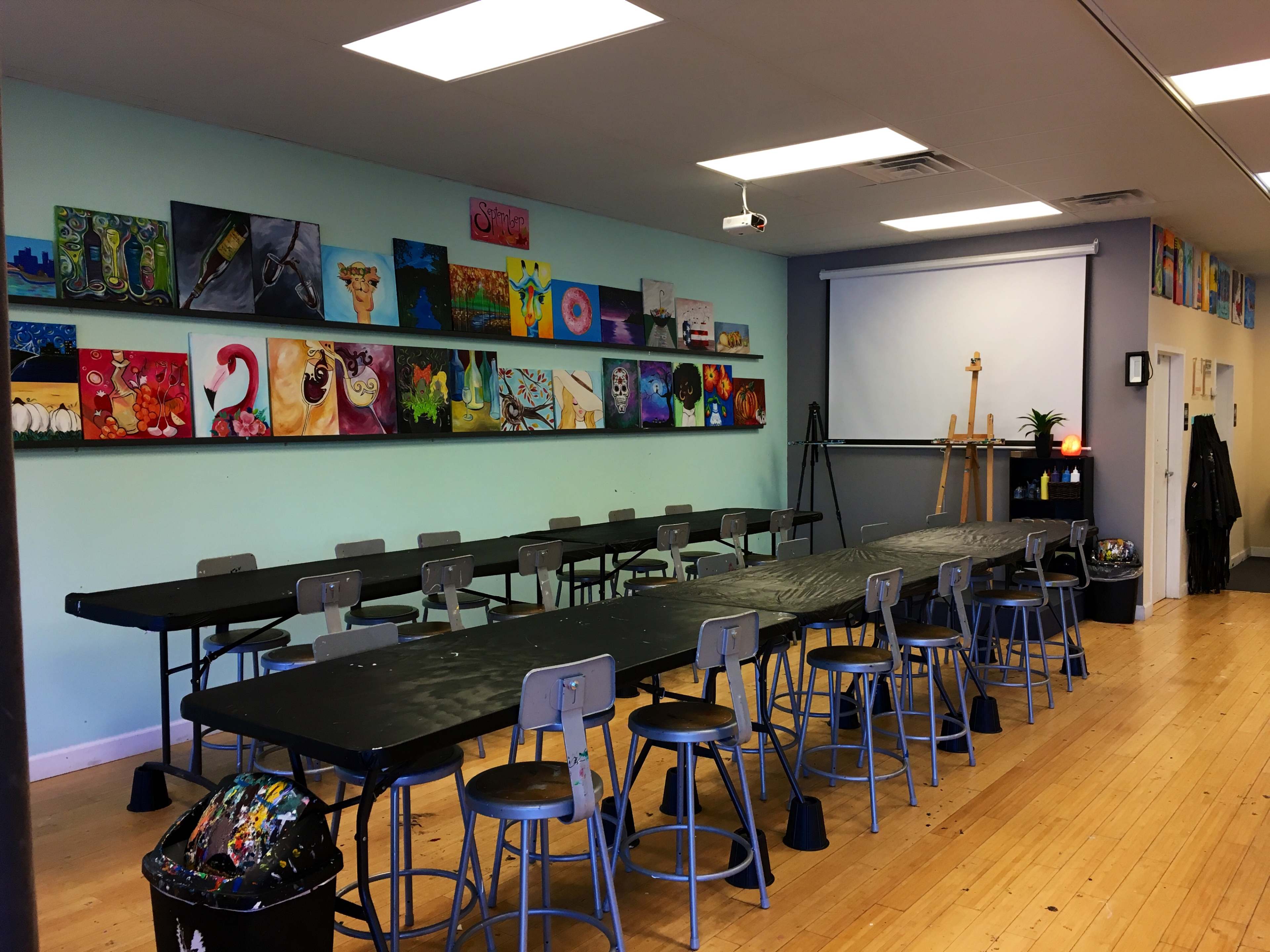 A spacious art classroom features long black tables and metal chairs, with colorful paintings displayed on the wall above and art supplies in the corner.