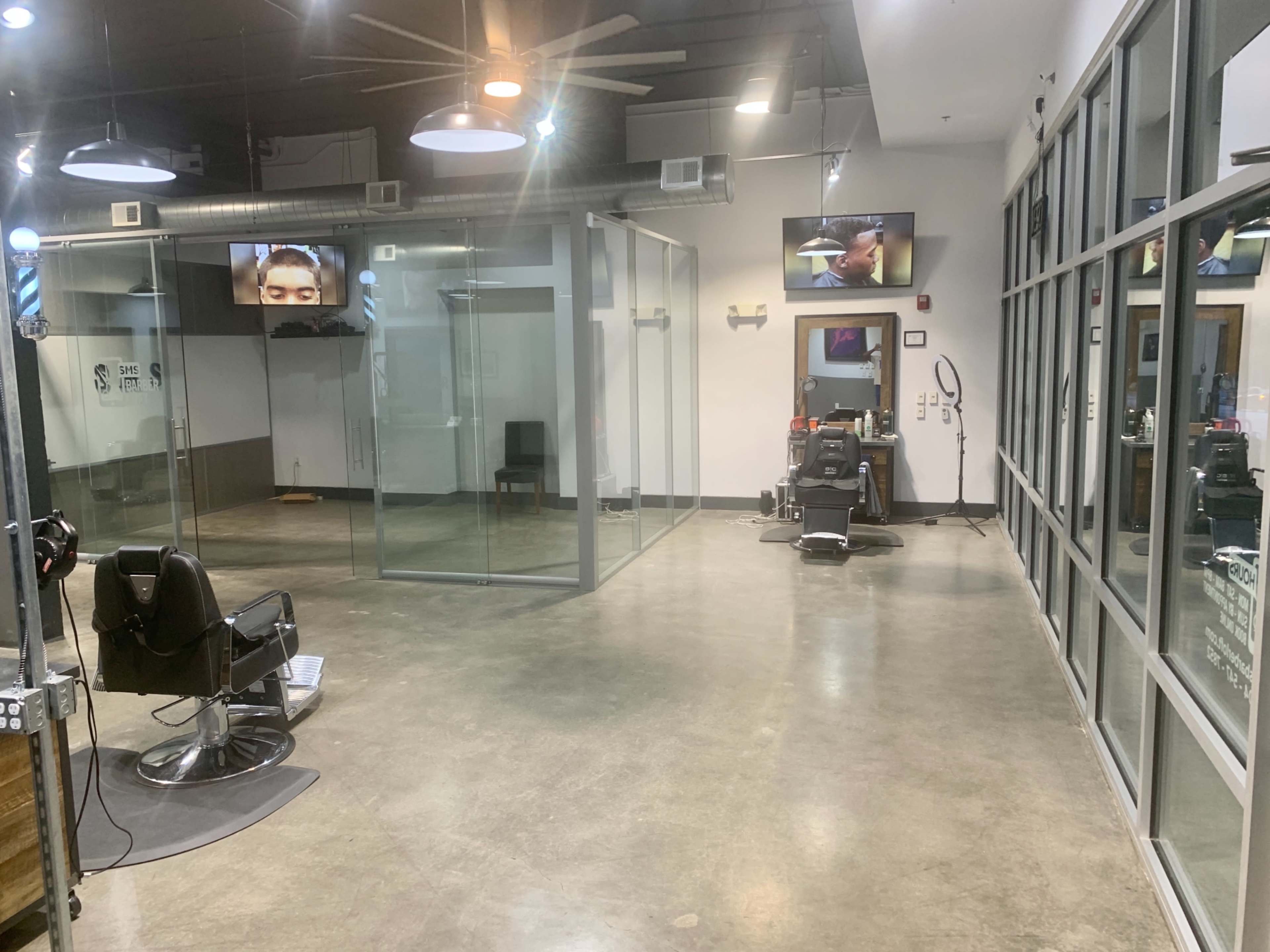The image shows a modern barbershop interior with a sleek design, featuring glass partitions, hair styling chairs, and two televisions mounted on the walls.