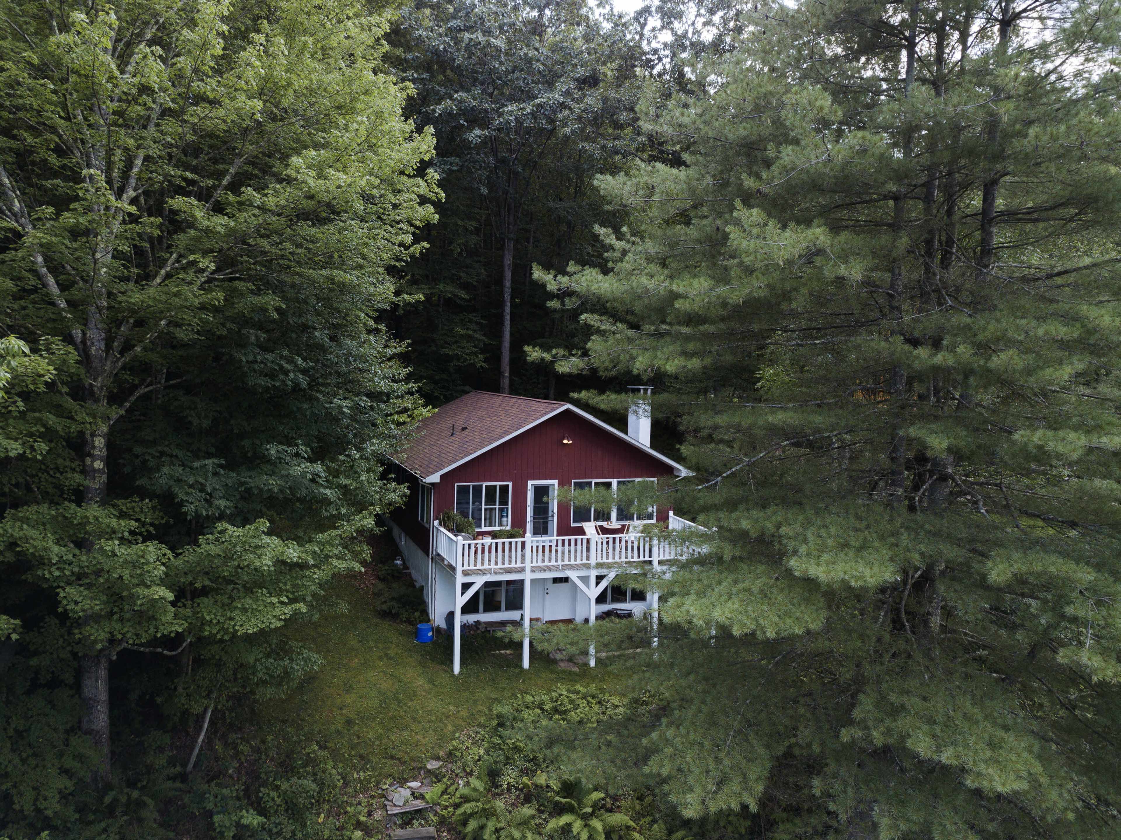 A small red cabin is nestled among dense trees, featuring a white deck and surrounded by greenery.