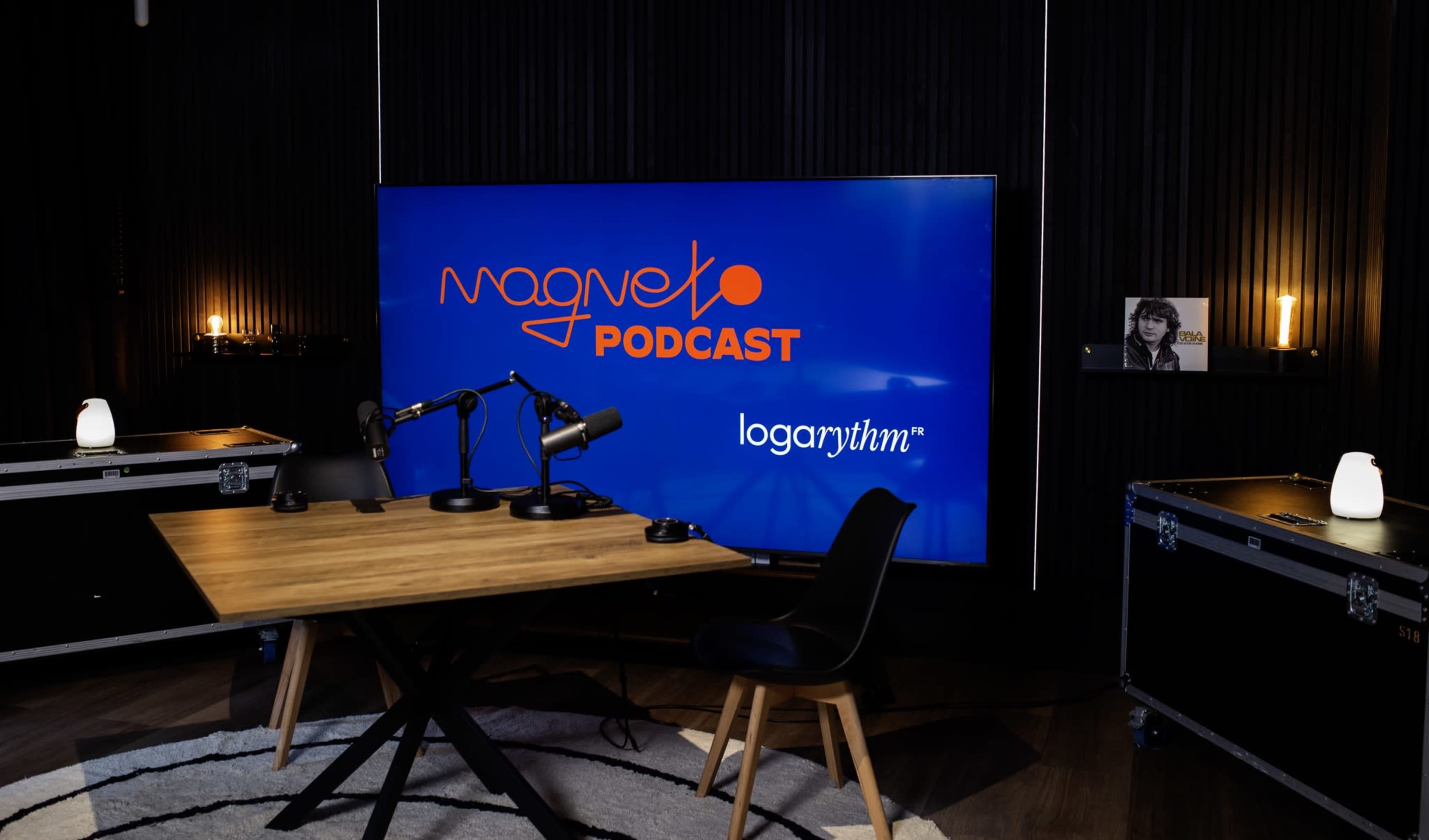 A podcast studio features a large screen displaying the logo "Magneto Podcast" alongside "logarythm," with a wooden table and microphones set up in front of it.