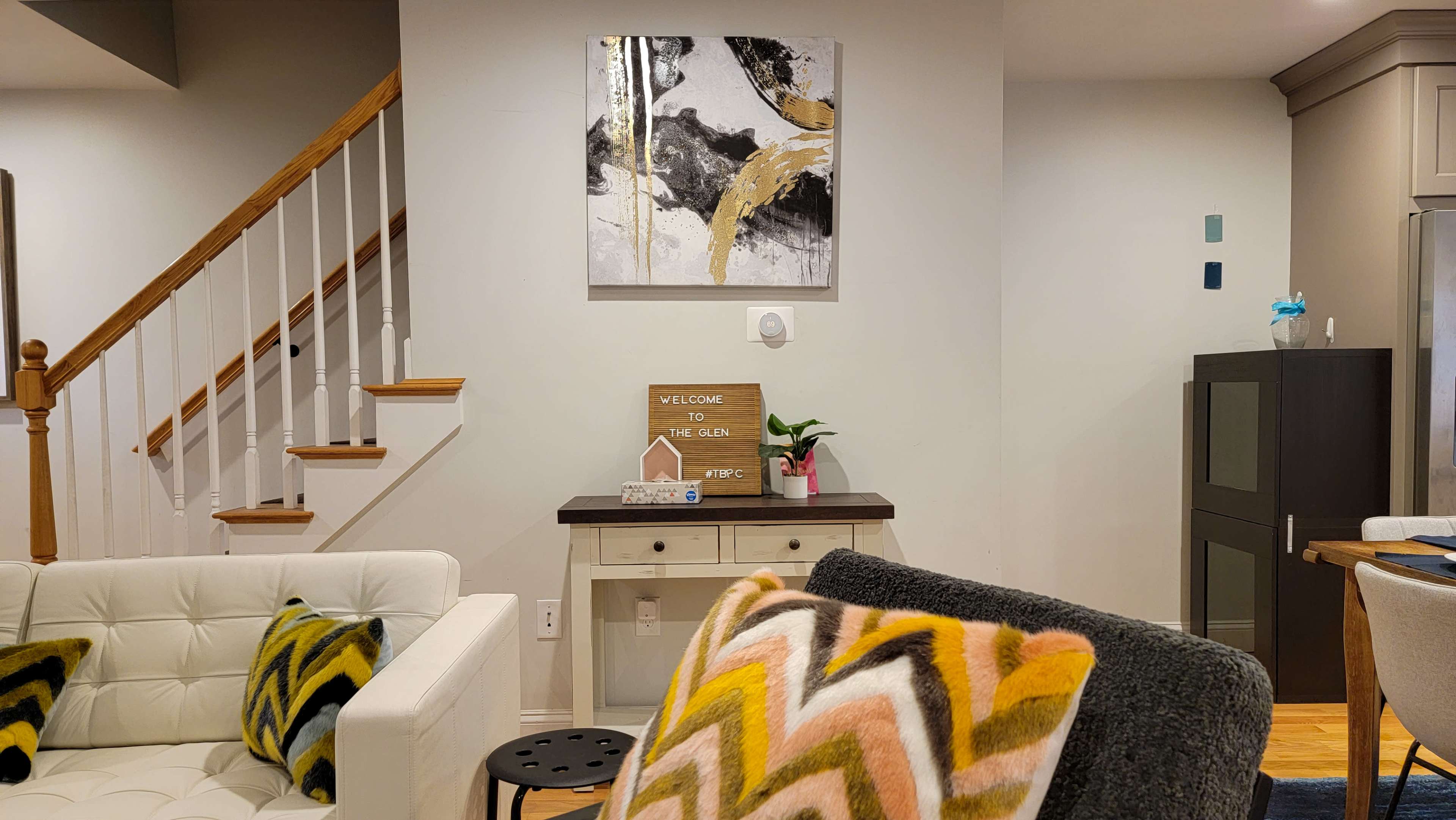 A cozy living room featuring a white couch, a small table with a welcome sign, a colorful abstract painting on the wall, and stairs leading to an upper level.