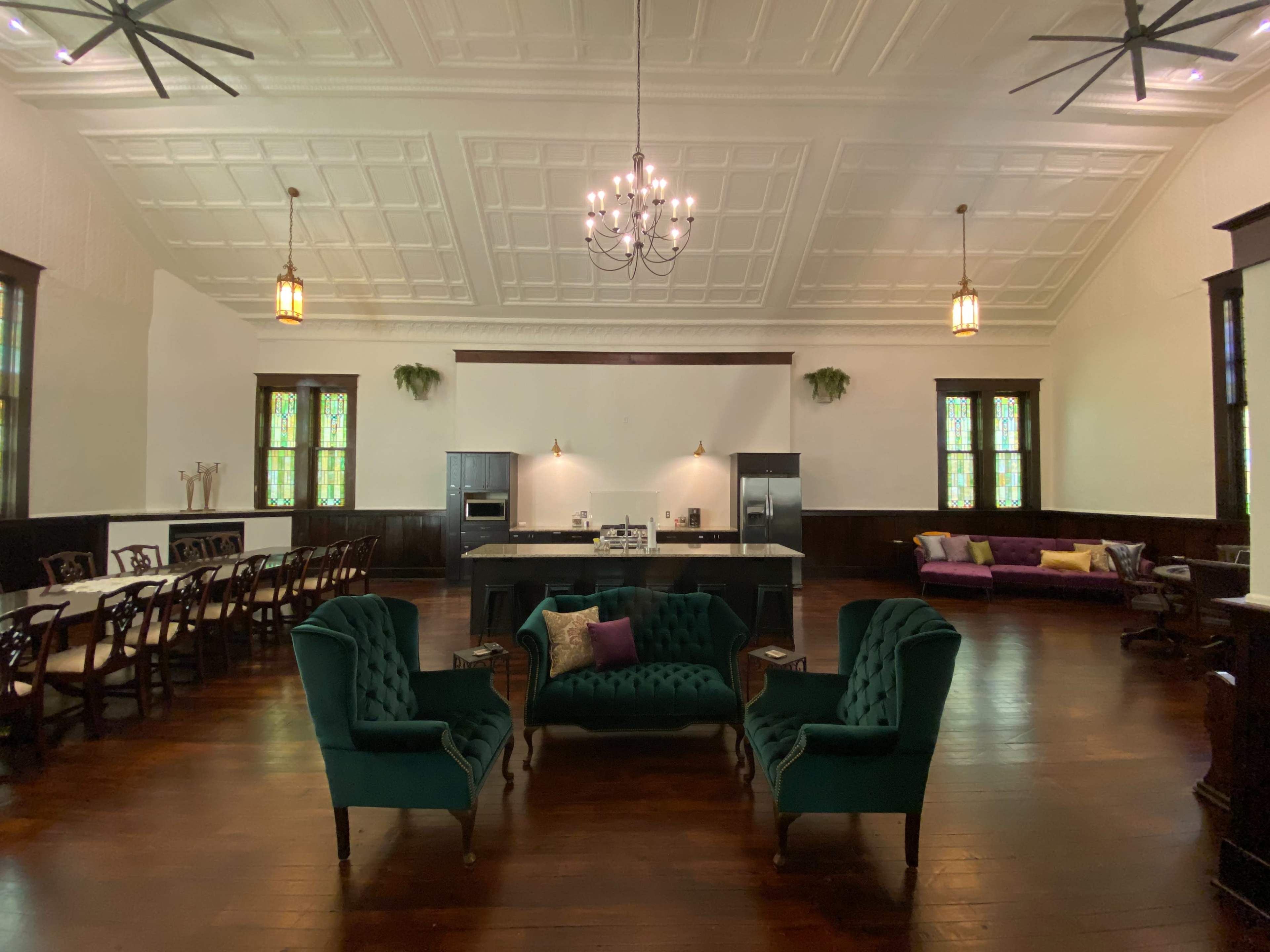 The image shows a spacious interior with a dining area featuring a long table and chairs, a kitchen area, and a seating arrangement of green velvet armchairs in the center.