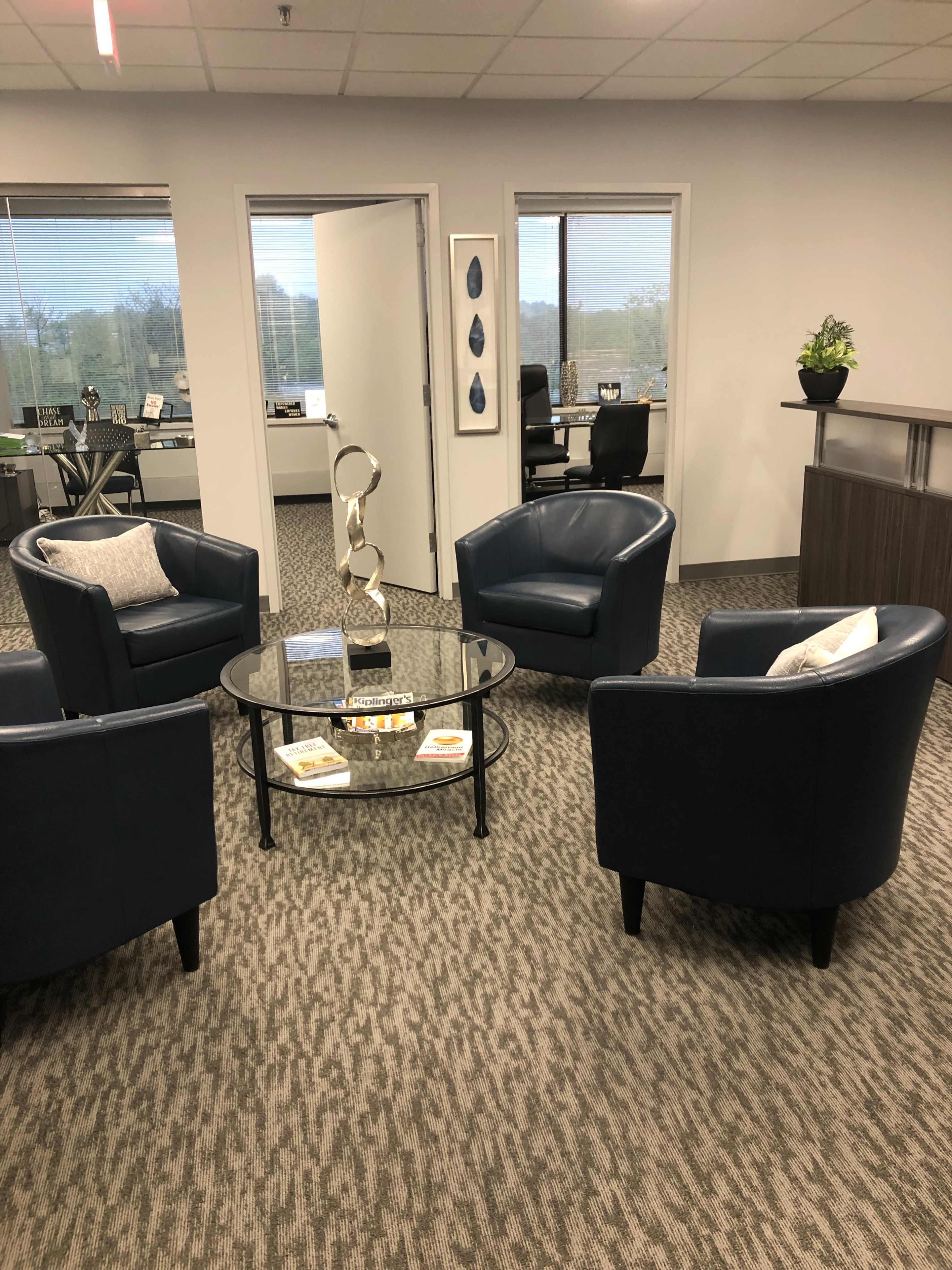 A modern office reception area with four blue armchairs arranged around a glass coffee table on a patterned carpet.