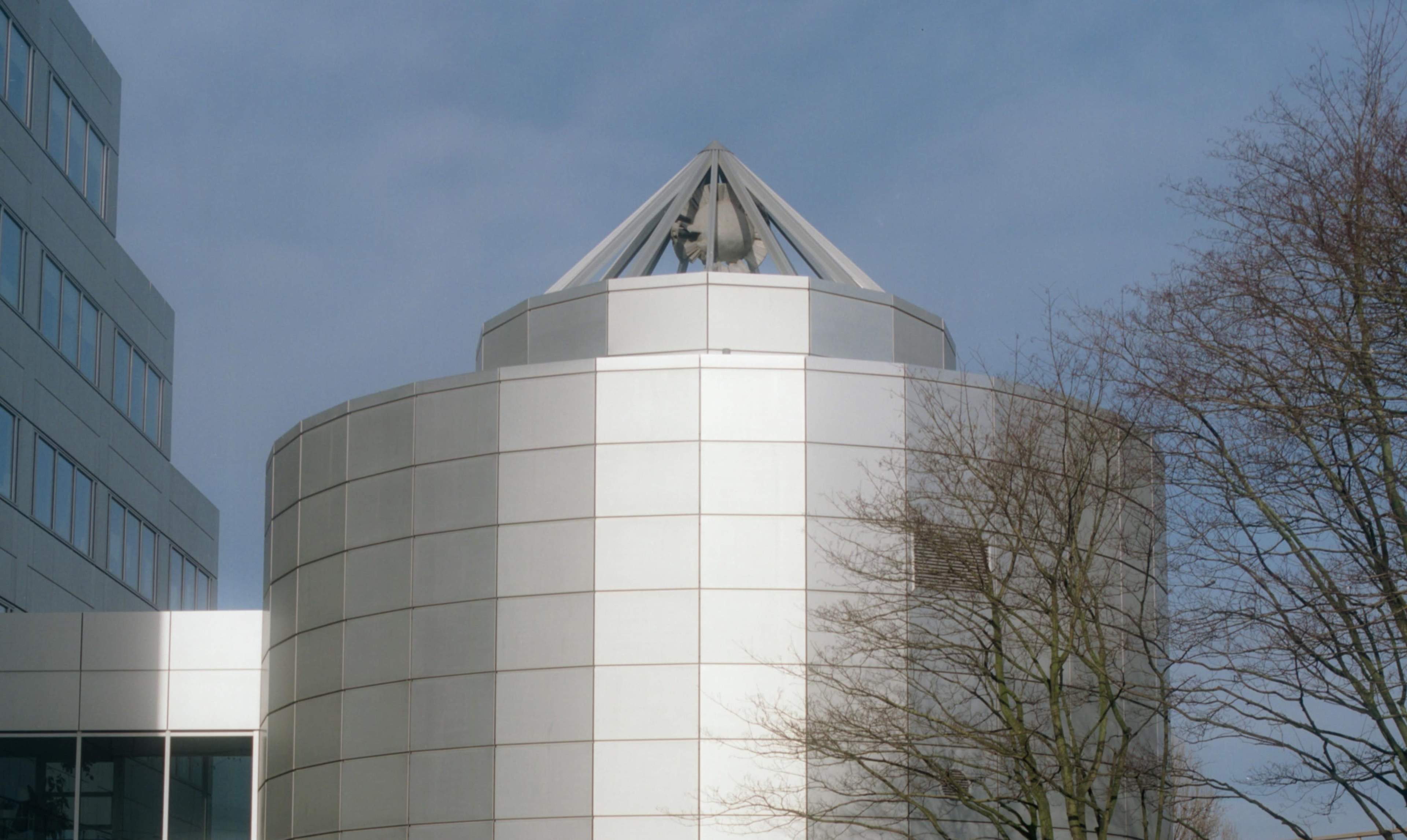 The image shows a modern cylindrical building topped with a triangular structure and surrounded by bare trees against a cloudy sky.