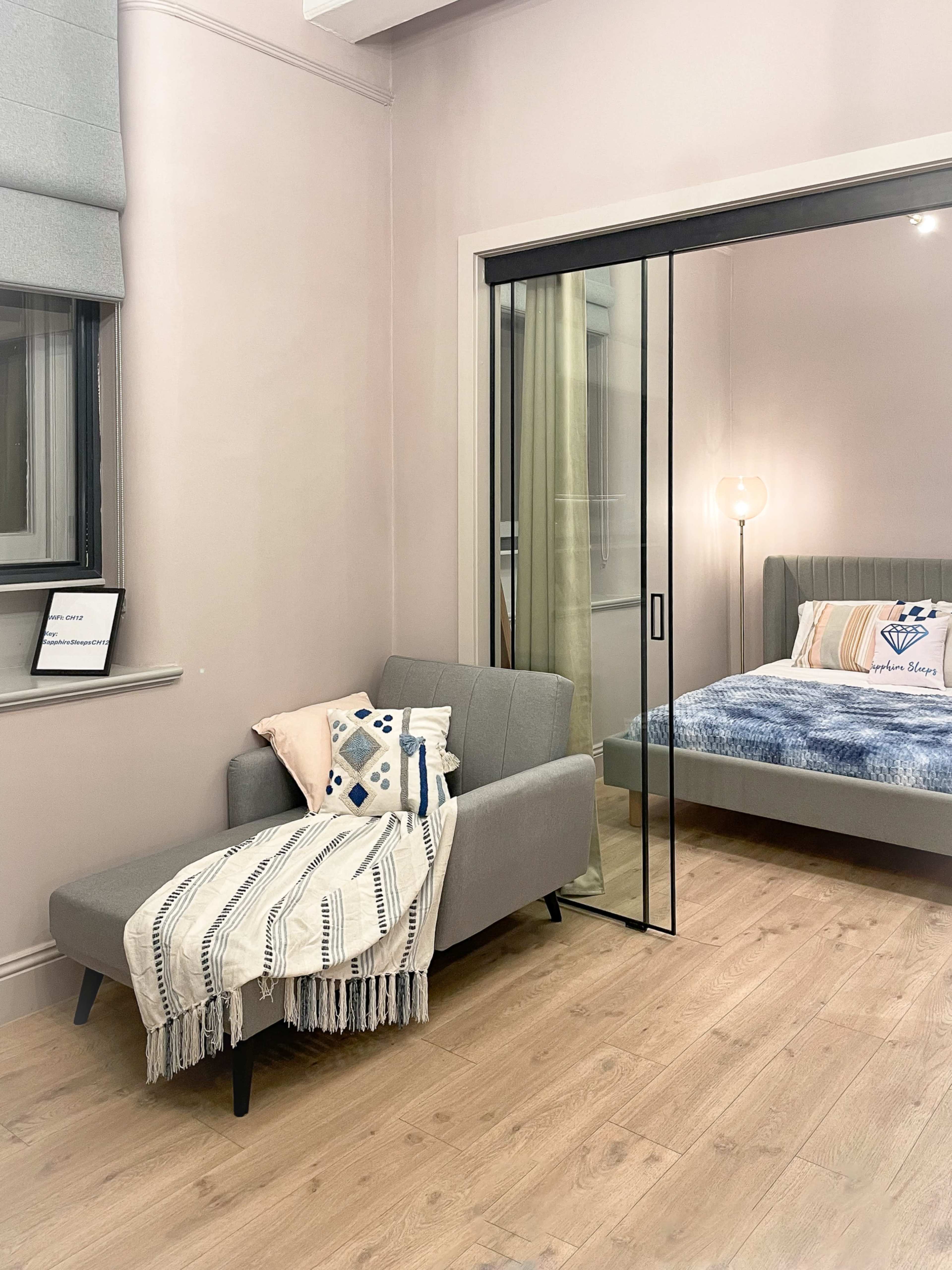 The image shows a small, modern bedroom with a gray sofa and a bed enclosed by a glass partition.