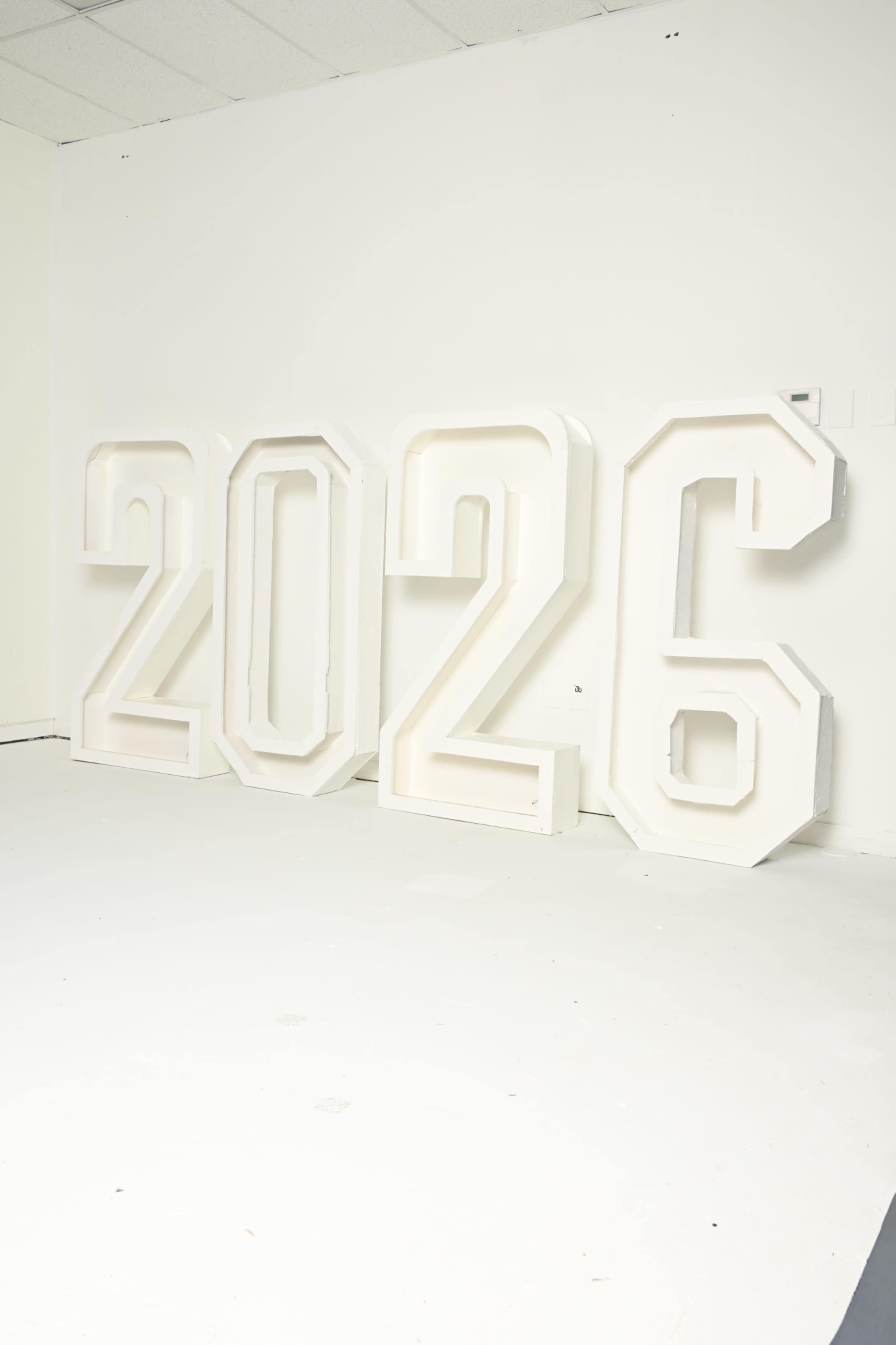 The image shows large, three-dimensional white numbers "2026" standing upright against a plain white wall.