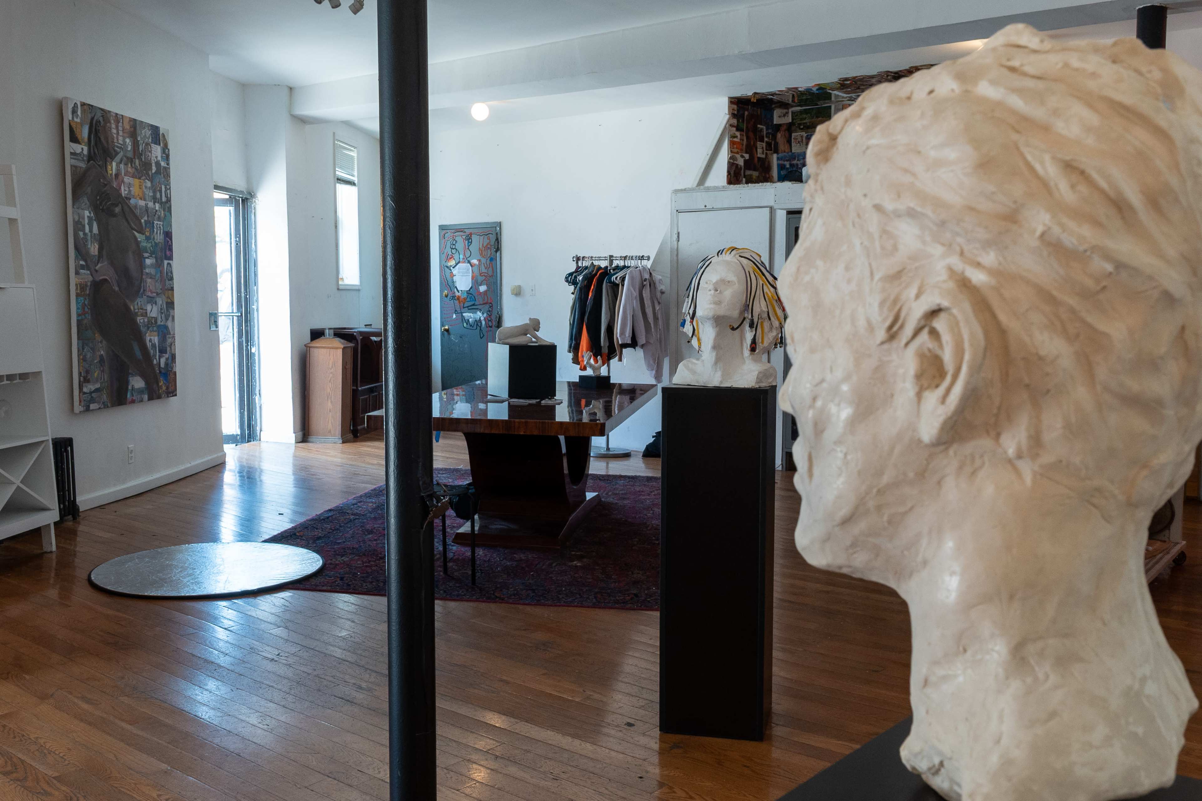 An art studio featuring a sculpture of a head in the foreground, with a wooden table and various artworks visible in the background.