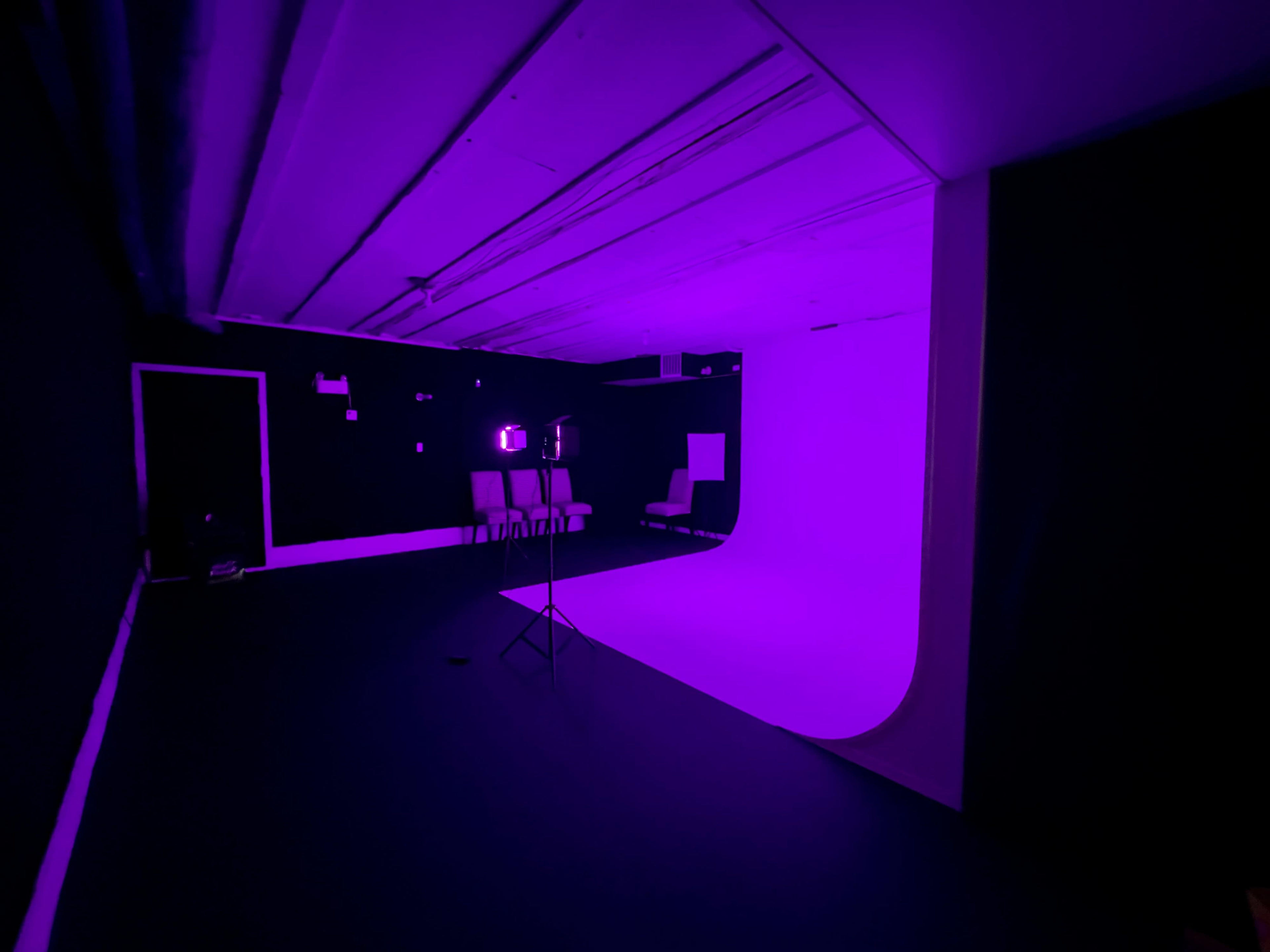 A studio space is illuminated in purple light, featuring a curved backdrop, a chair, and a tripod stand in the foreground.